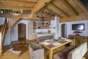 Un vaste espace de vie agrémenté d’un escalier en bois, d’une table à manger avec des chaises rembourrées, d’un lustre et d’une cuisine située au fond de la pièce.