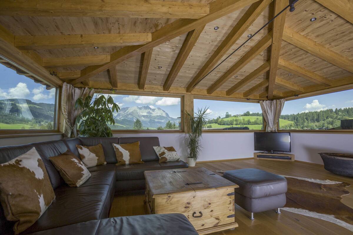 Ein geräumiges Wohnzimmer mit Holzbalkendecke, einem großen Fenster mit Blick auf die Berge, einem Ledersofa, einem Couchtisch und einem Fernseher.