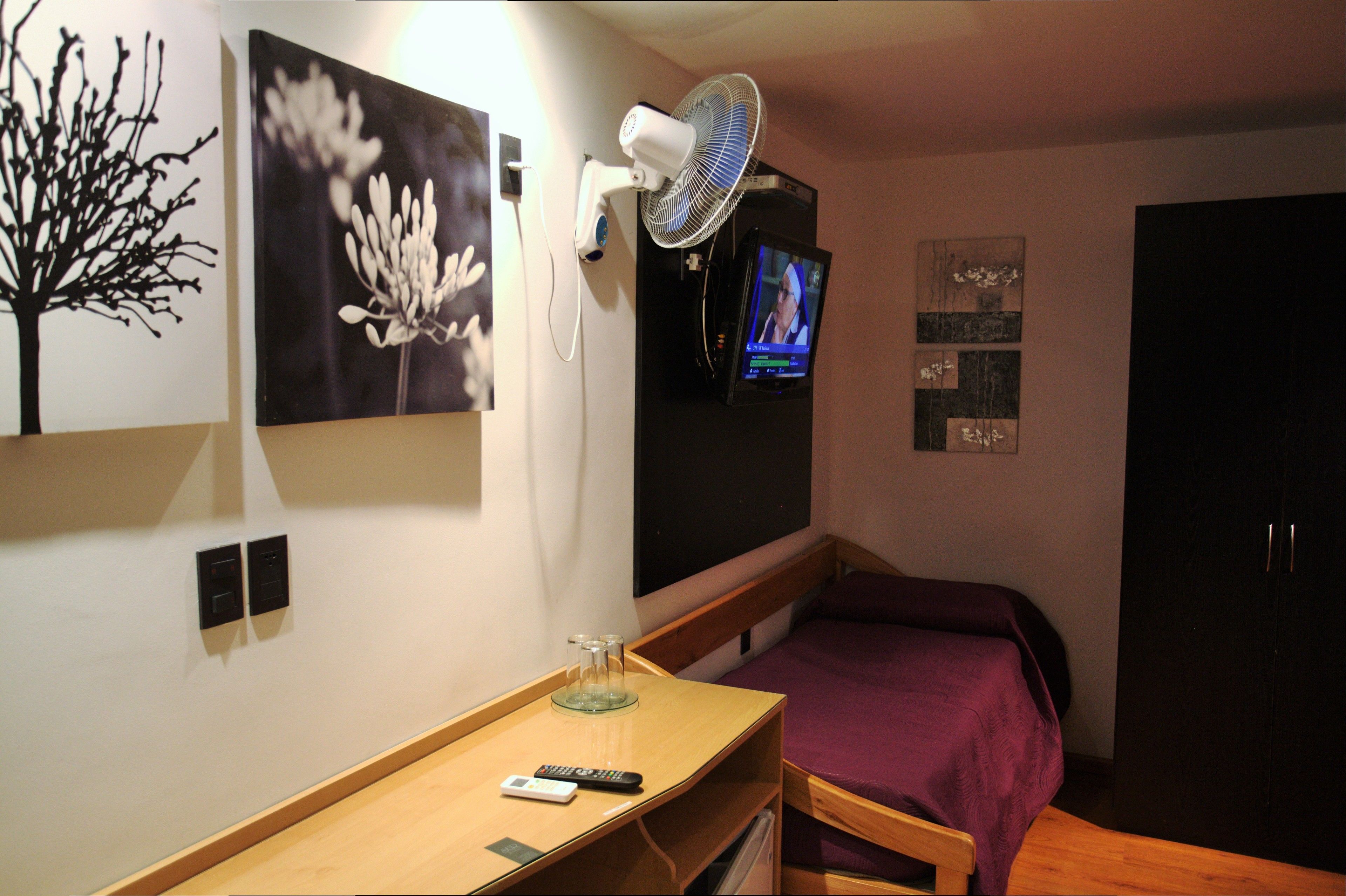 Una habitación de hotel con una cama, un escritorio con un control remoto, un televisor montado en la pared y un ventilador de pared.
