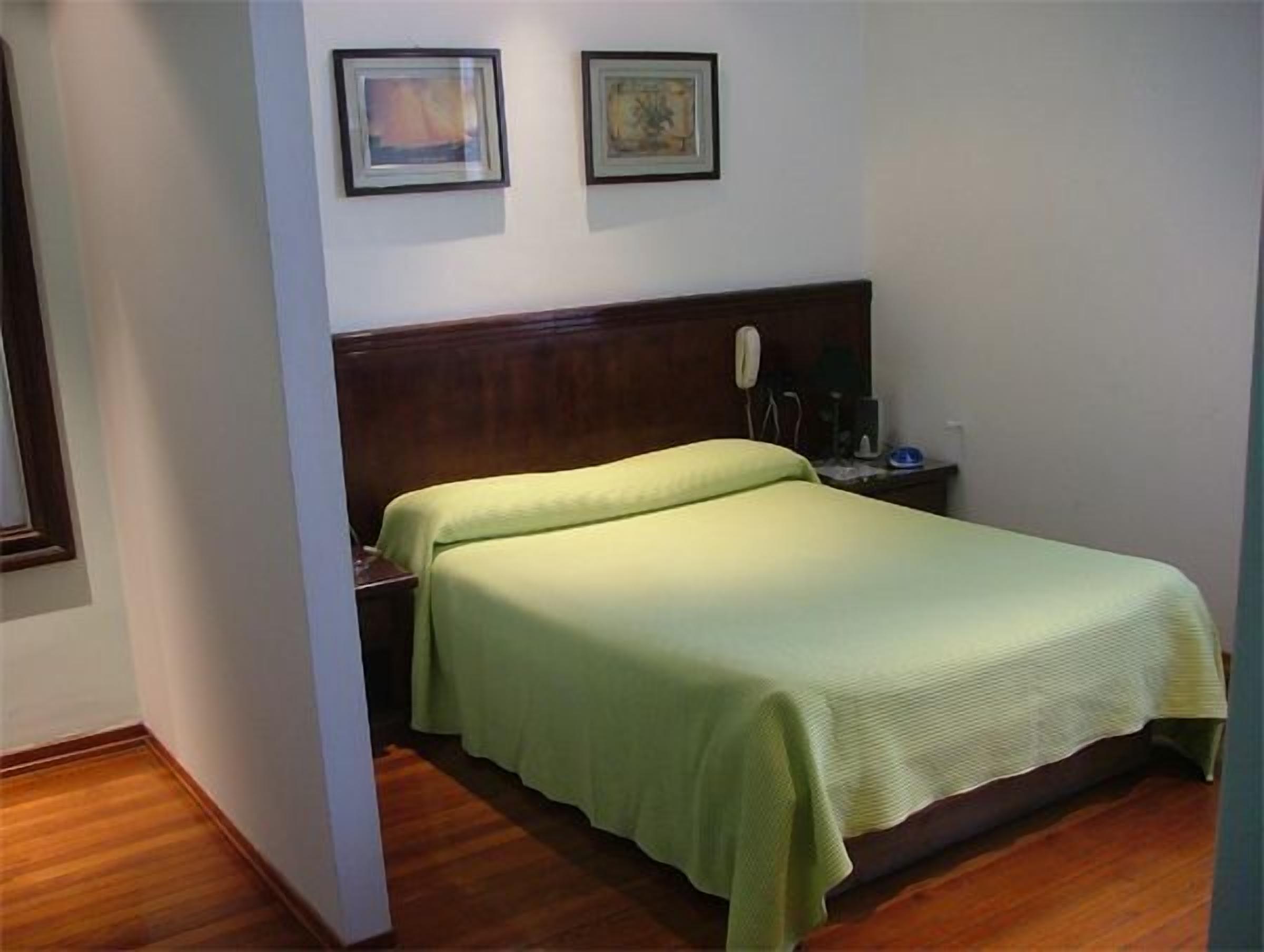 Una cama individual con cabecera de madera, una mesita de noche con lámpara y dos cuadros enmarcados en la pared.