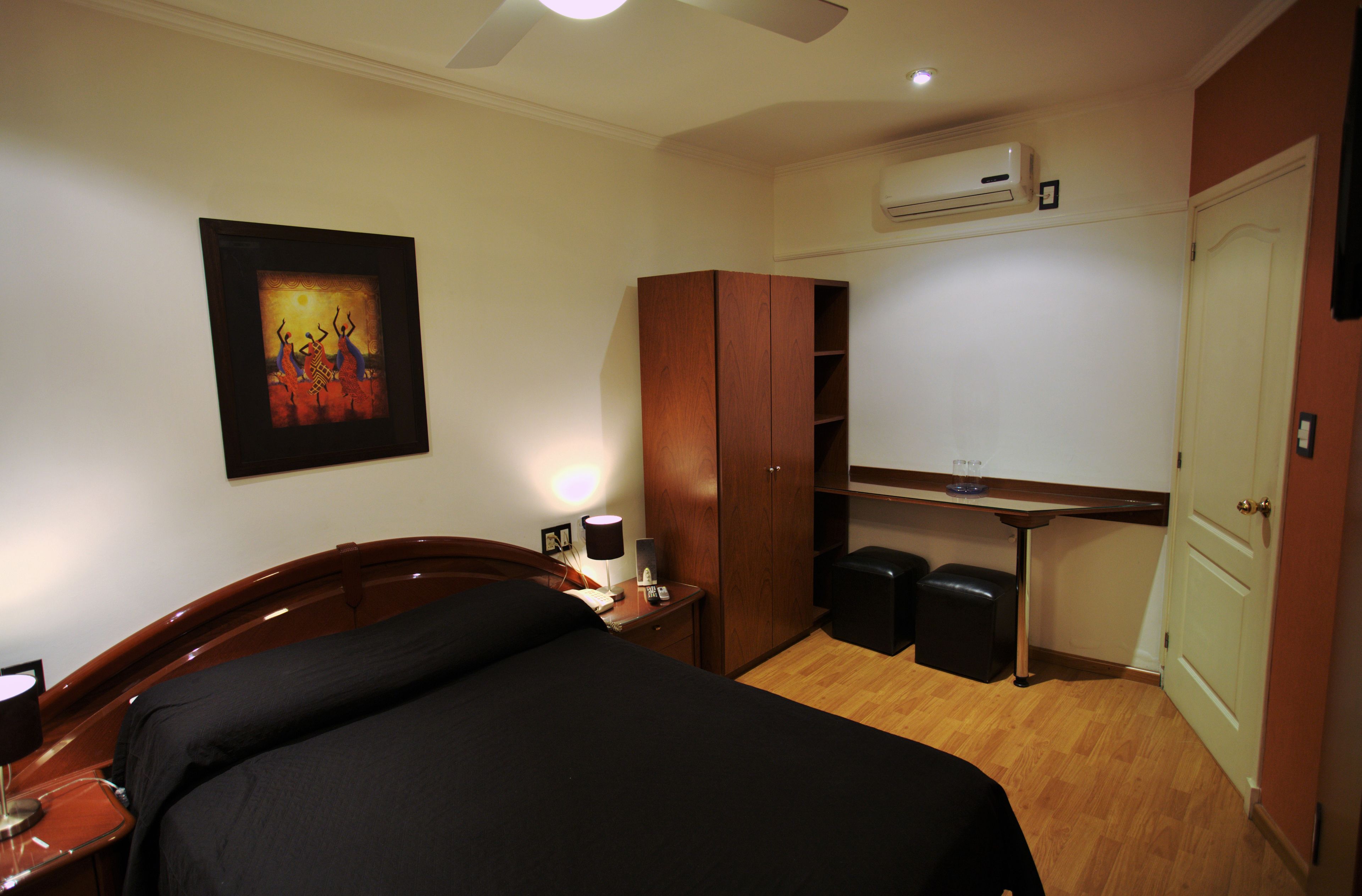 A hotel room with a bed, a desk, a wardrobe, a trash bin, and an air conditioning unit.