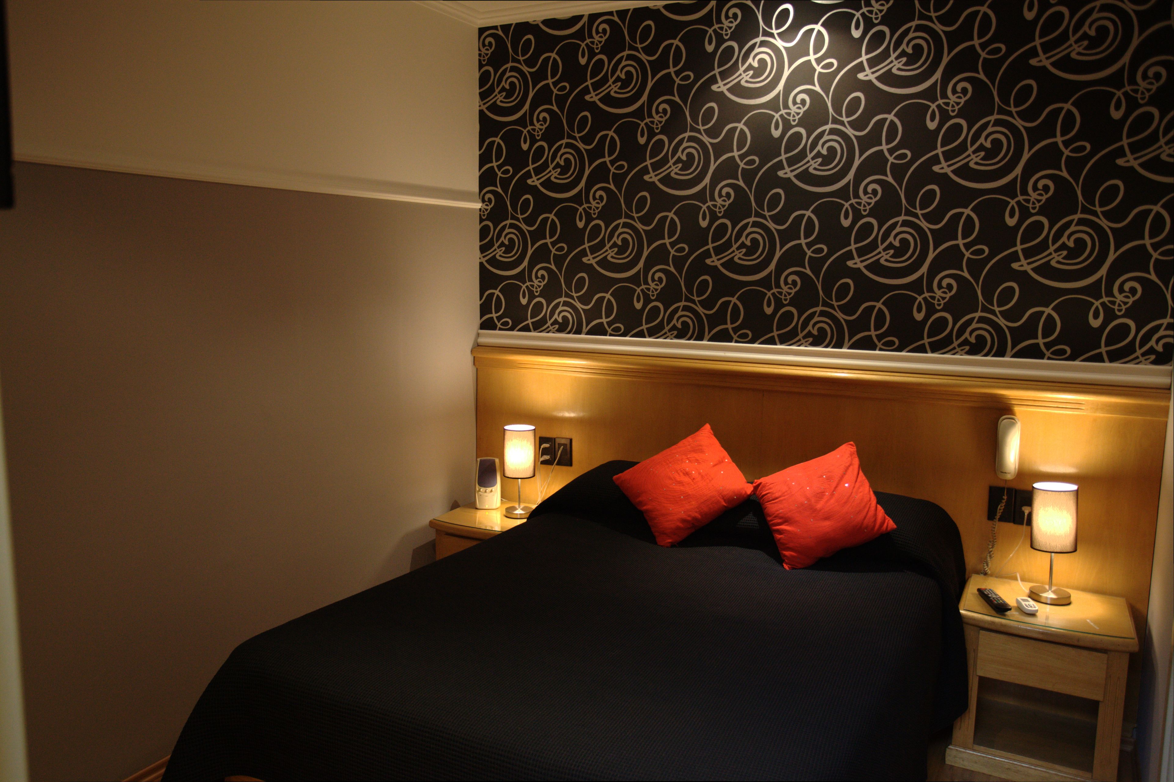 Una habitación de hotel con una cama, dos mesitas de noche con lámparas, un teléfono y un papel tapiz estampado.