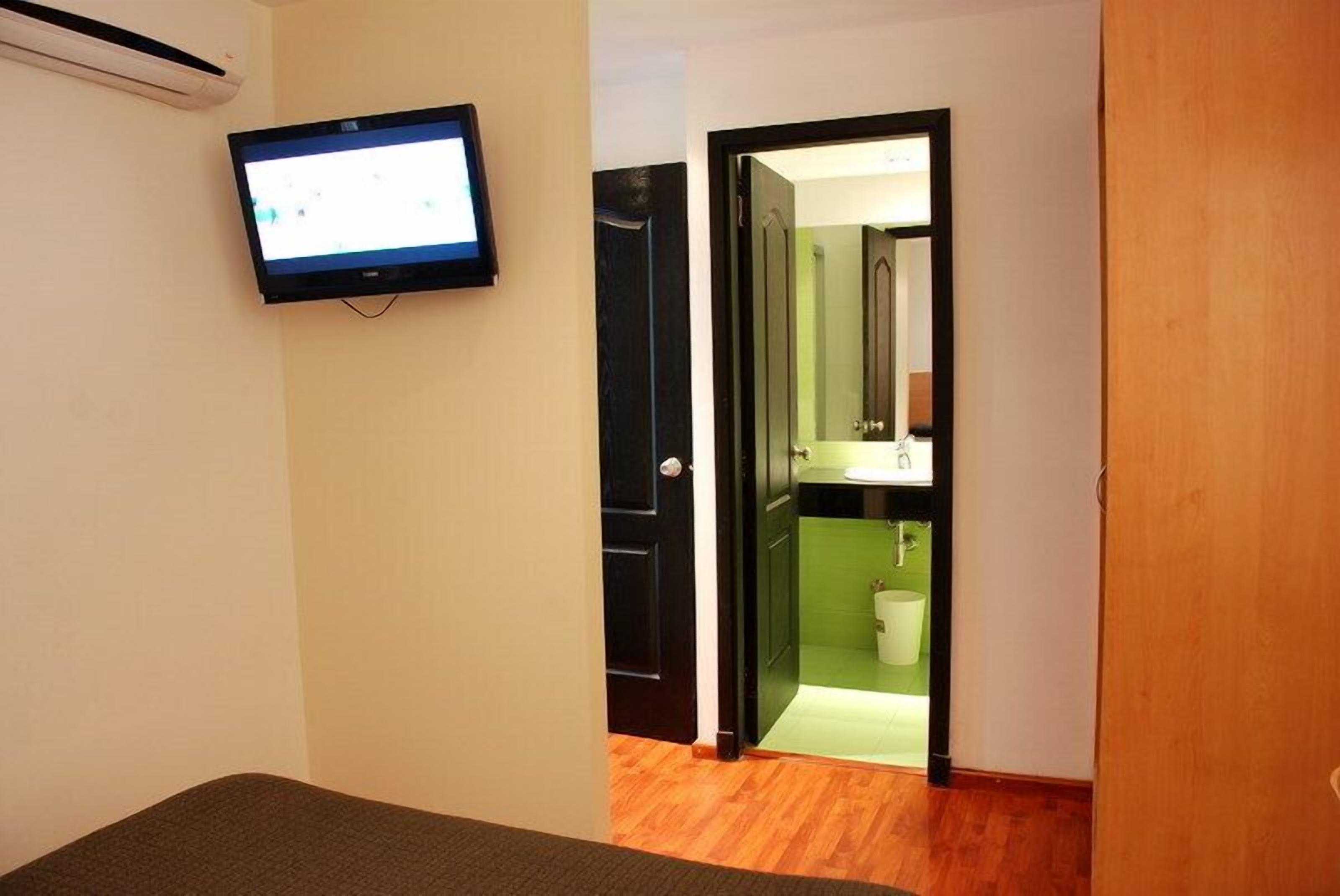 Una habitación con cama, televisor montado y puerta que da a un baño con lavamanos de azulejos verdes.