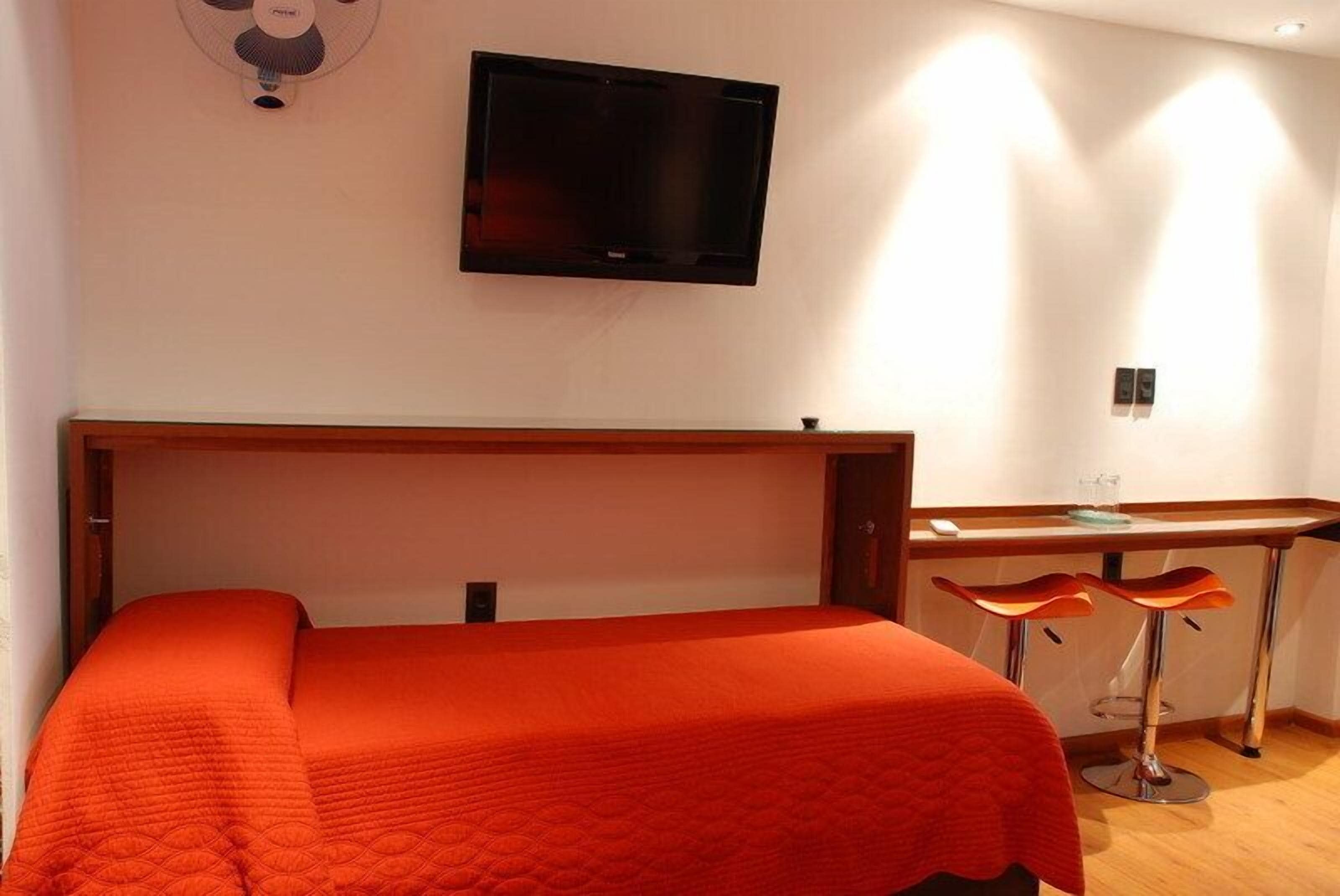 Una habitación de hotel con una cama, un televisor montado en la pared y un escritorio con dos taburetes.