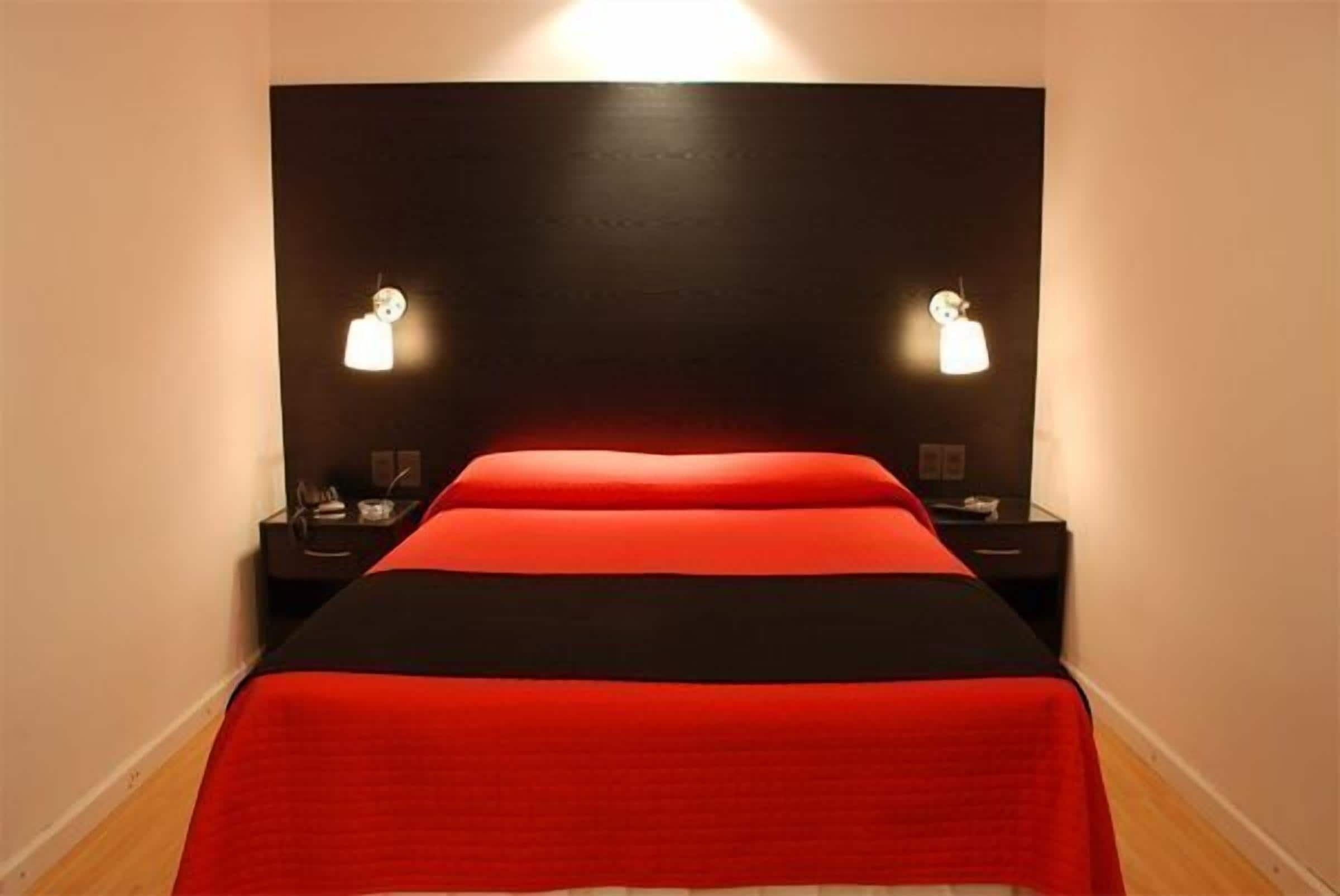 Una cama con cabecera negra y ropa de cama roja, dos lámparas de noche y una mesita con una lámpara.