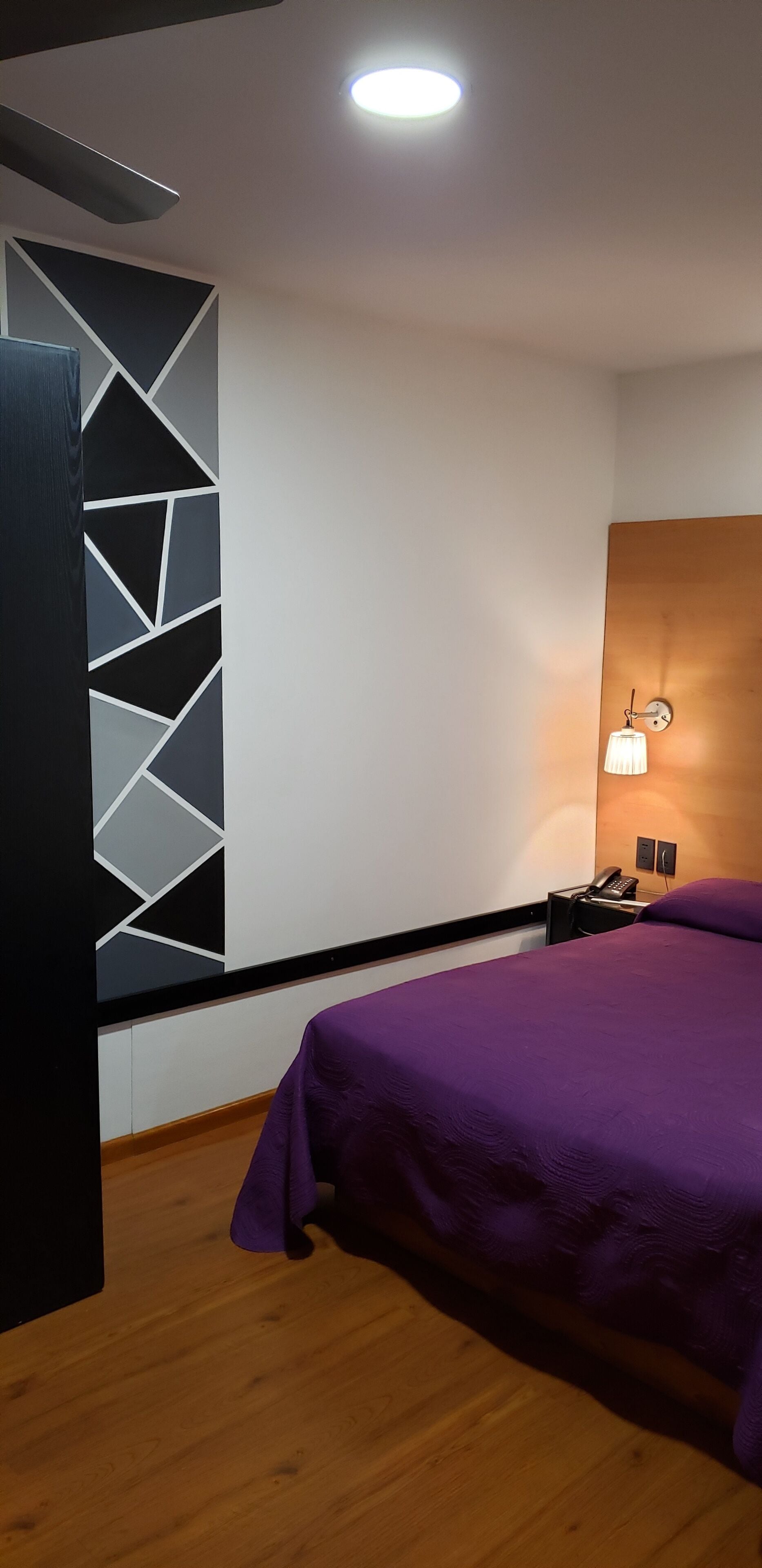 Una habitación de hotel moderna con una cama morada, arte mural geométrico, cabecero de madera, mesita de noche con lámpara y ventilador de techo.