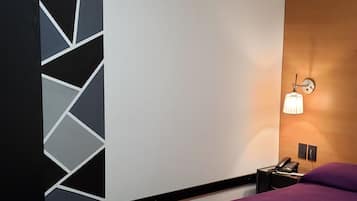 Una habitación de hotel moderna con una cama morada, arte mural geométrico, cabecero de madera, mesita de noche con lámpara y ventilador de techo.