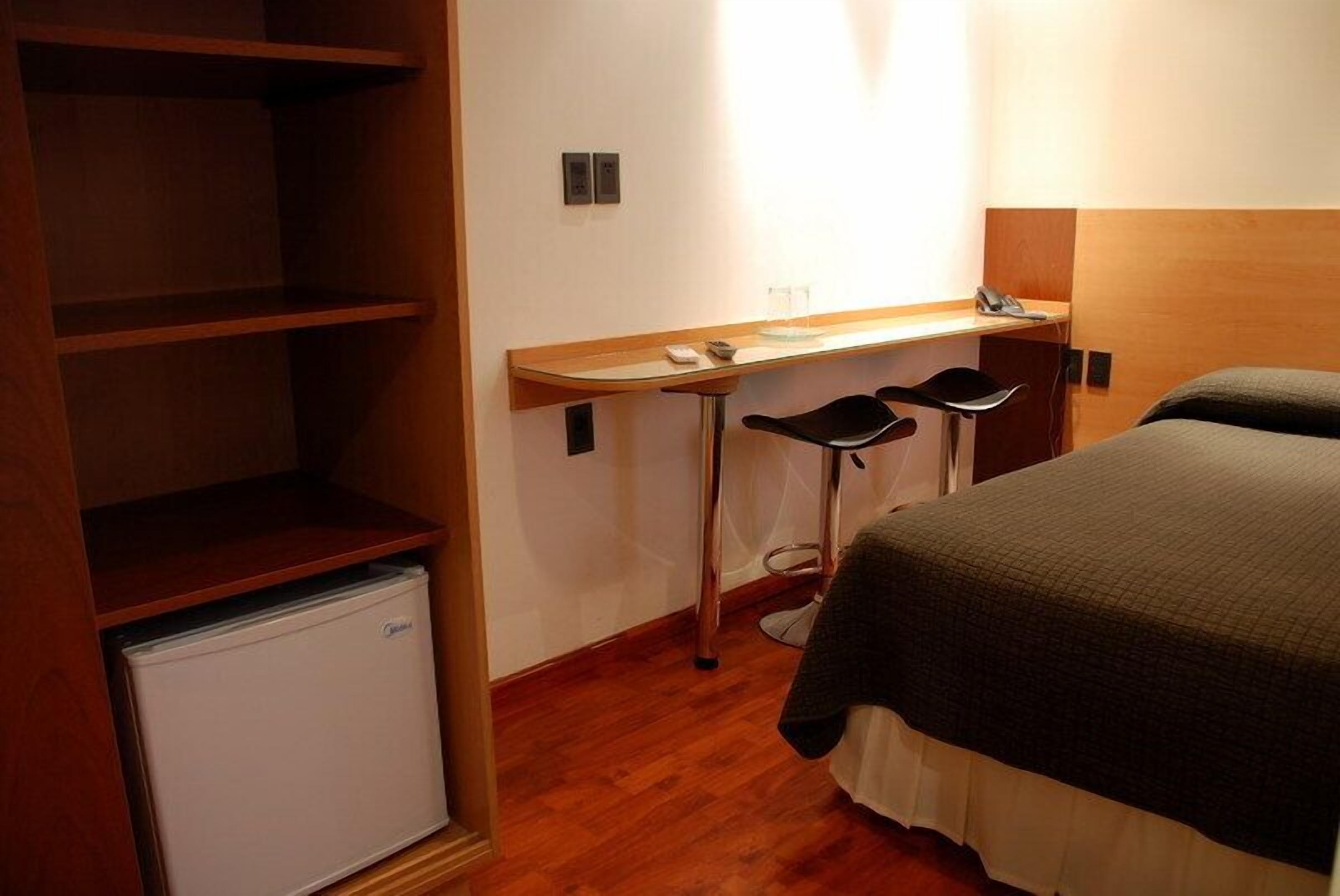 Ein kompaktes Hotelzimmer mit einem Bett, einem kleinen Schreibtisch mit zwei Stühlen, einer Minibar und einem Holzschrank.
