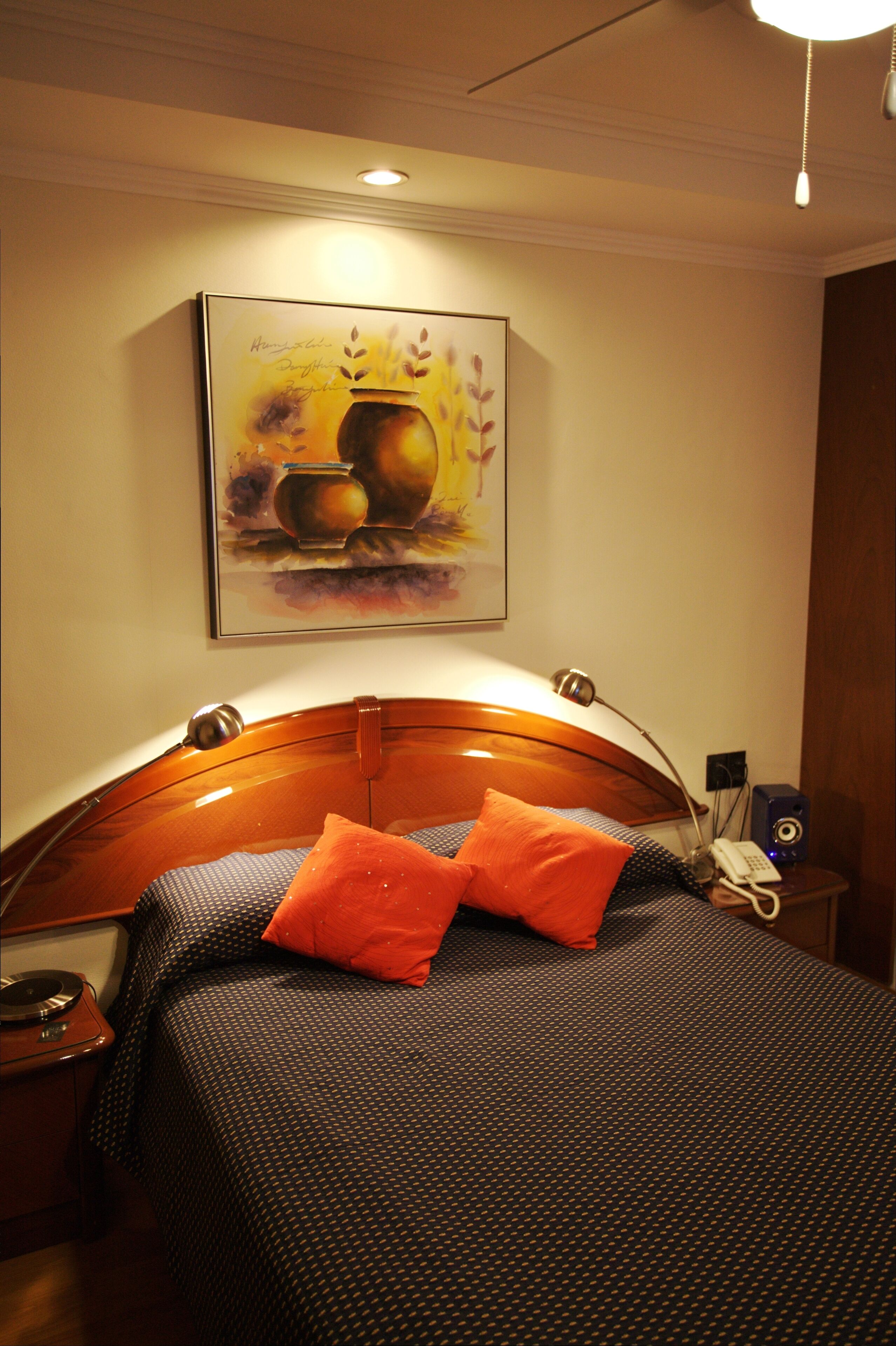 Habitación de hotel con cabecera de madera, cama con ropa de cama estampada, almohada naranja, un cuadro de frutas en la pared y una mesita de noche con una lámpara y un teléfono.