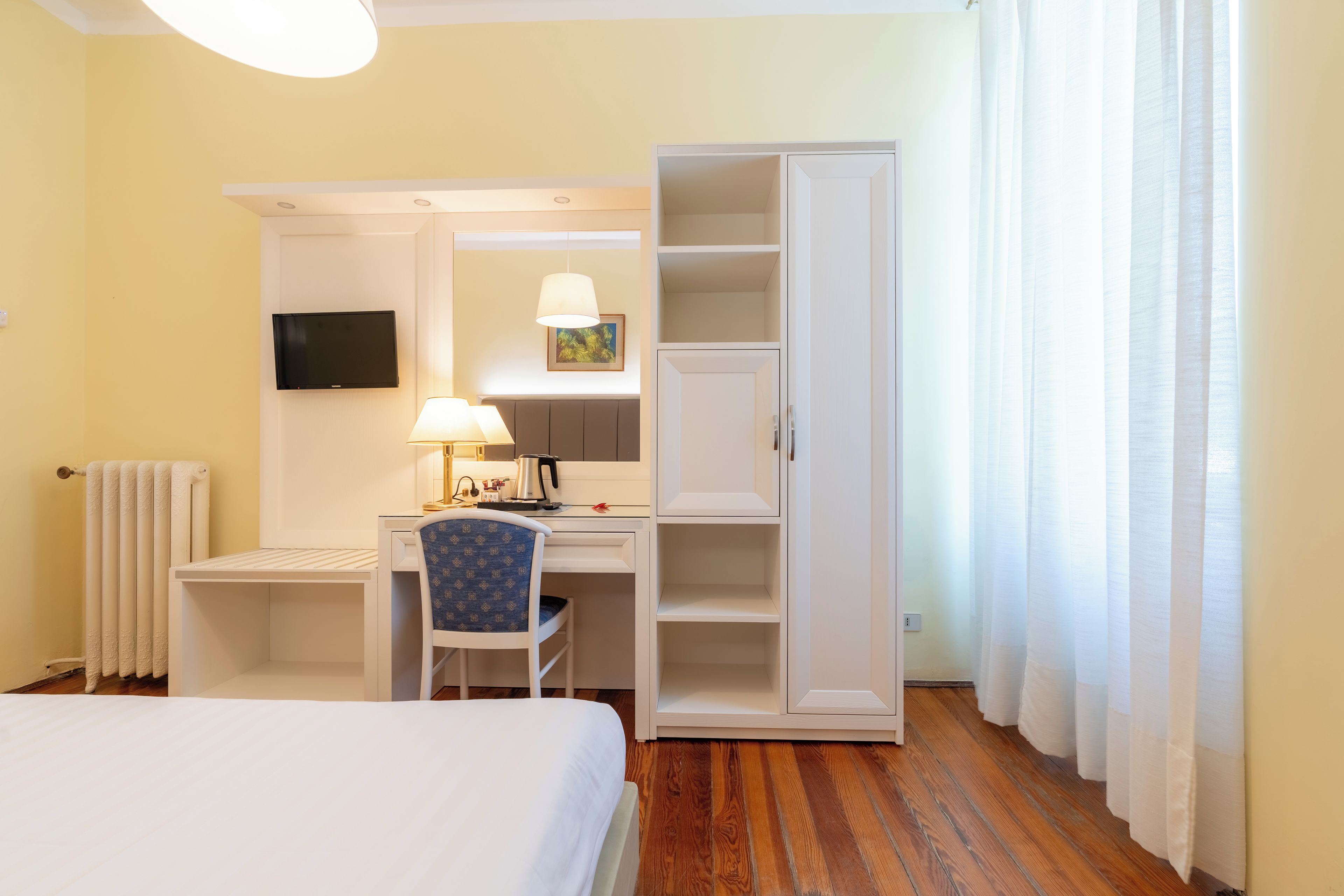 A hotel room with a bed, a desk with a chair, a television, a radiator, and a wardrobe.