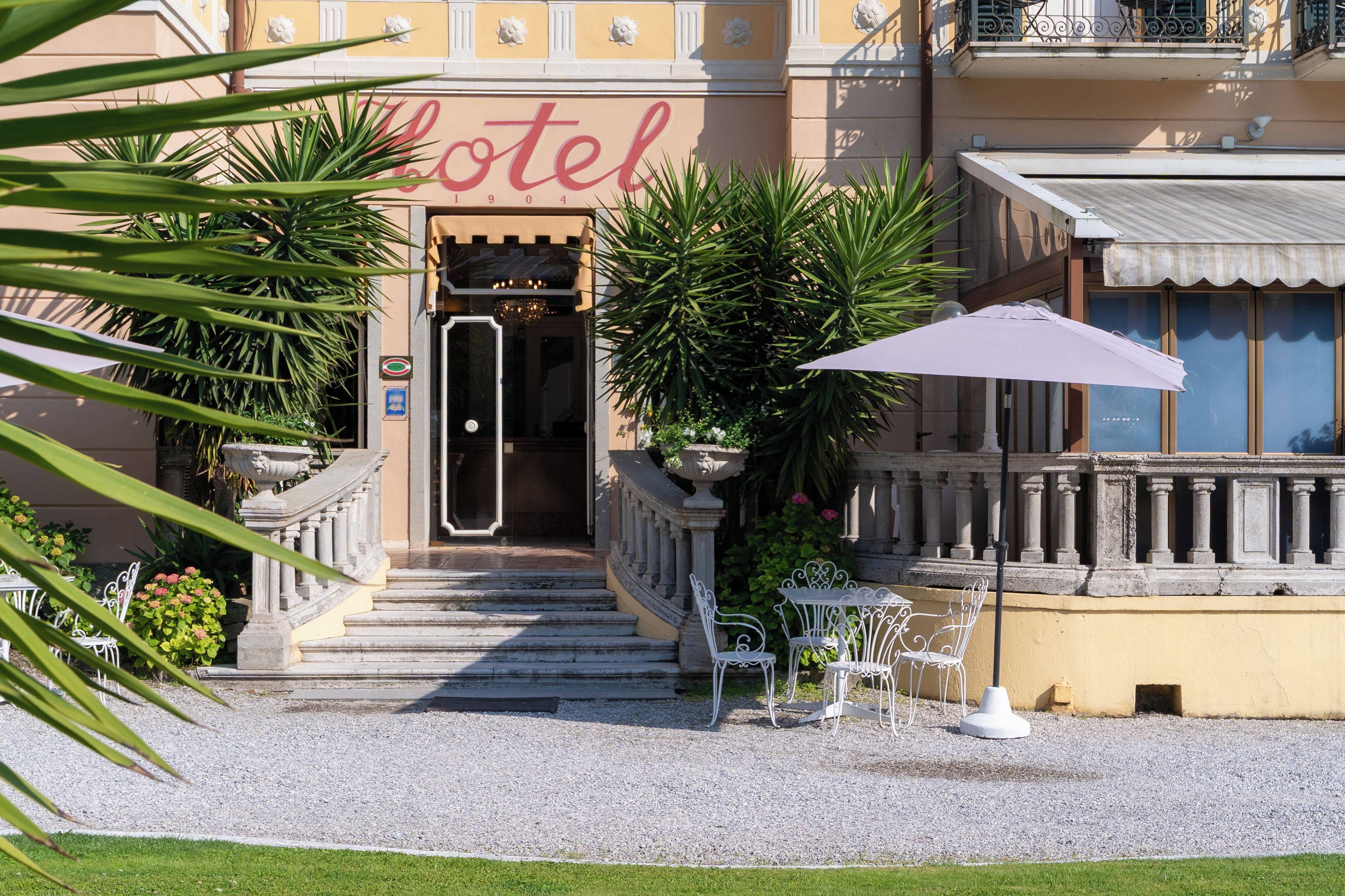 Ein Hotel mit Eingangsbereich, einer Treppe zum Eingang, einer überdachten Außenlounge und einem Garten mit Pflanzen.