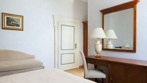 A hotel room with a bed, a desk with a lamp, a chair, and a door with a blue frame.