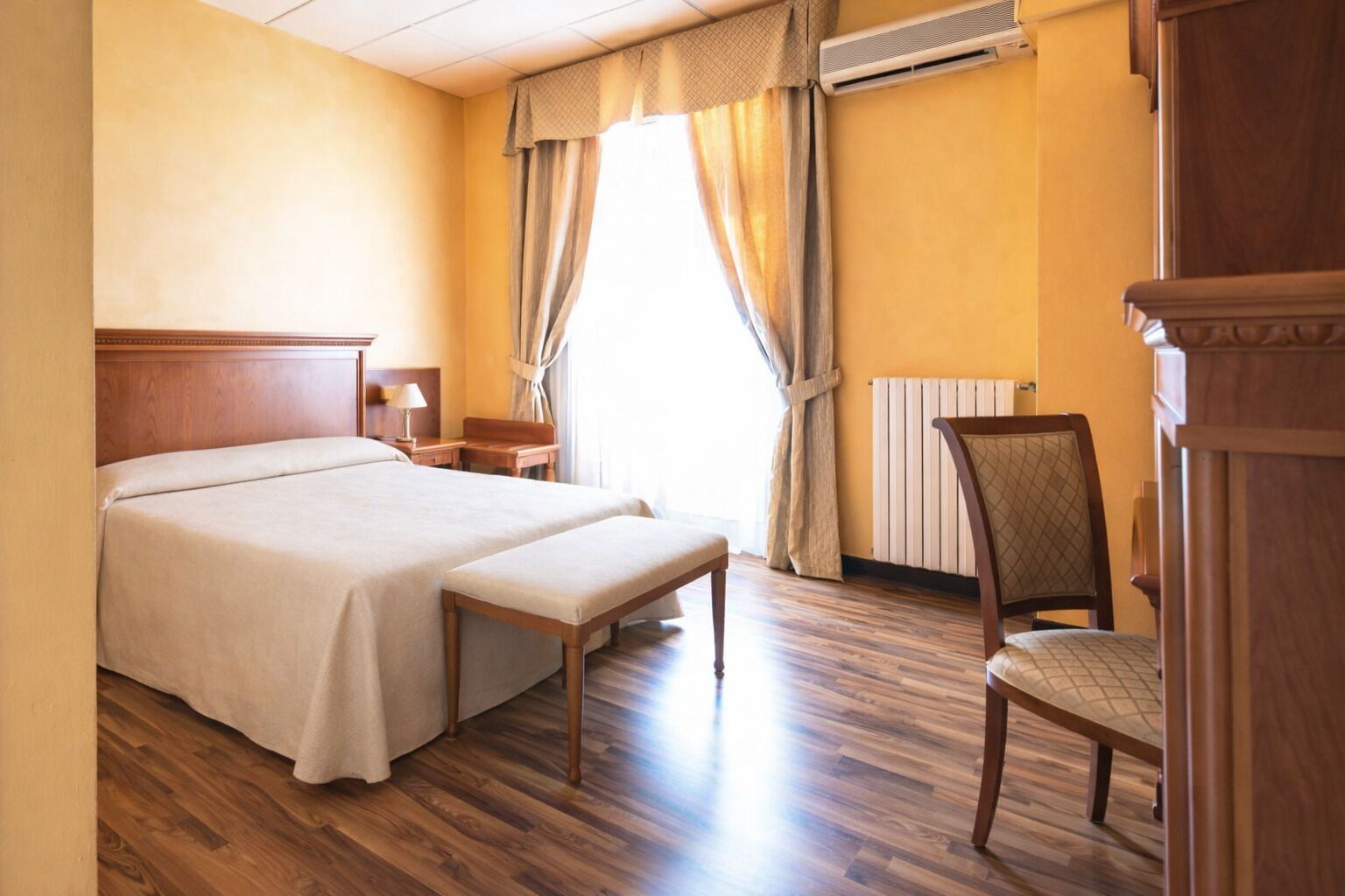 Une chambre d’hôtel avec un lit double, une table de chevet, une chaise, une fenêtre avec des rideaux et un radiateur.