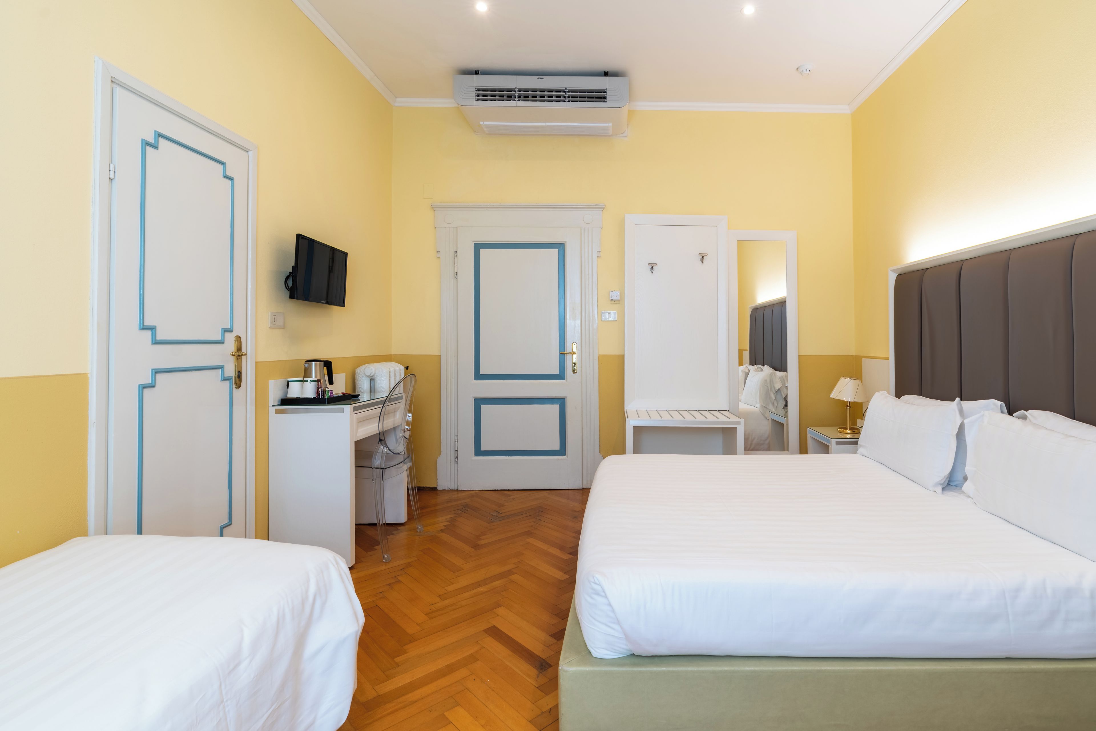 Habitación de hotel con dos camas, un escritorio con computadora, un espejo, un televisor montado en la pared y un equipo de aire acondicionado.