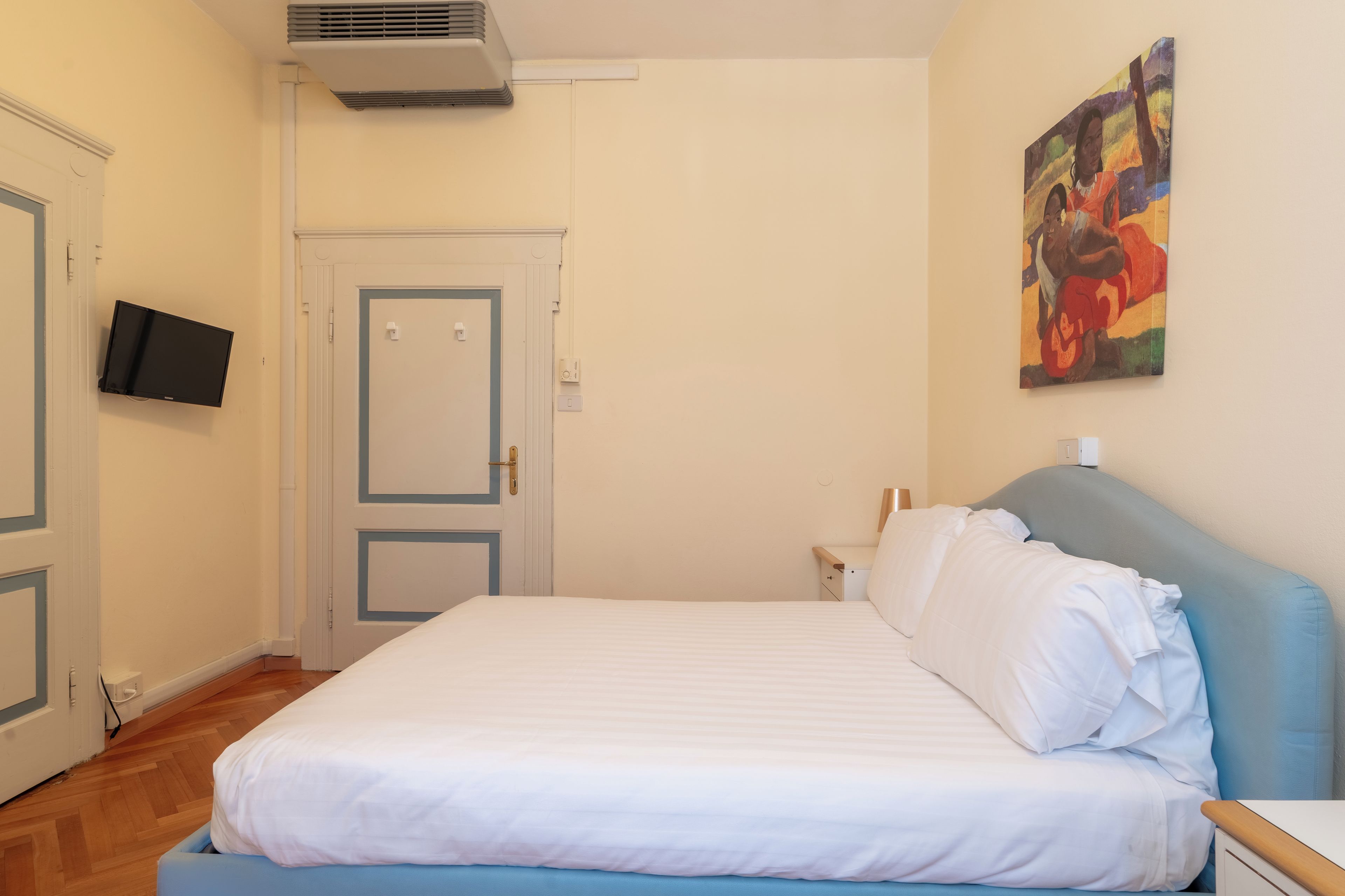 A bedroom with a bed, a flat-screen TV mounted on the wall, a door, and a painting.