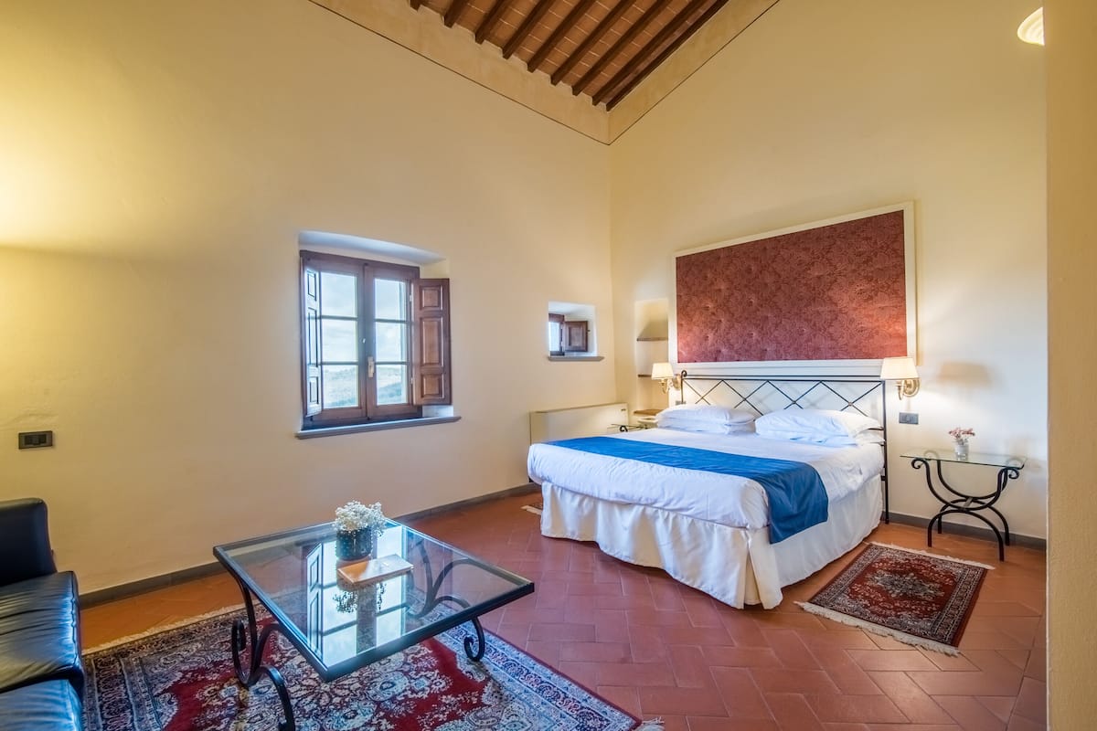 A bedroom with a bed, a glass table with a vase, a window, and a red rug.