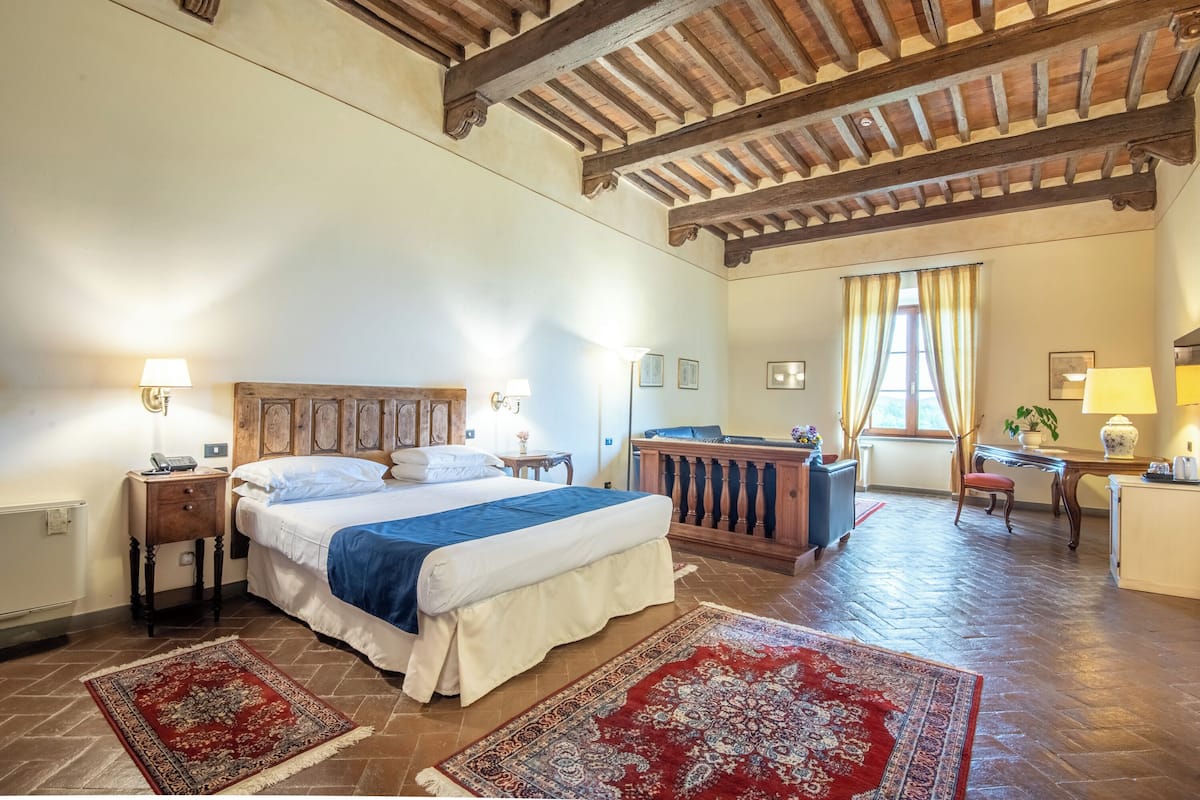 A bedroom with a wooden headboard, a large bed, two bedside tables with lamps, a wooden bench, a desk with a chair, and a window with curtains.