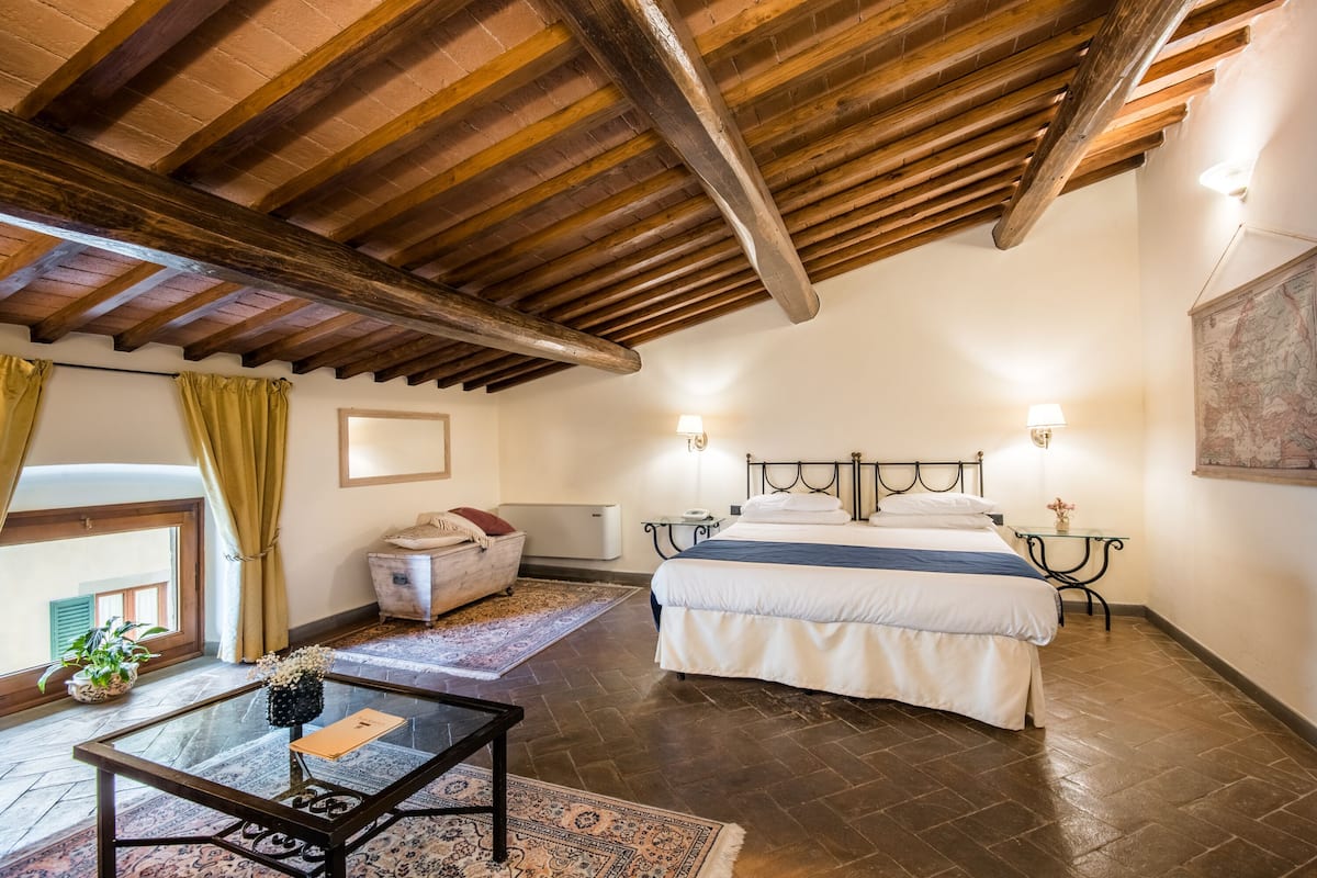 A bedroom with a large bed, a small table, a chair, a nightstand, and a window with curtains.
