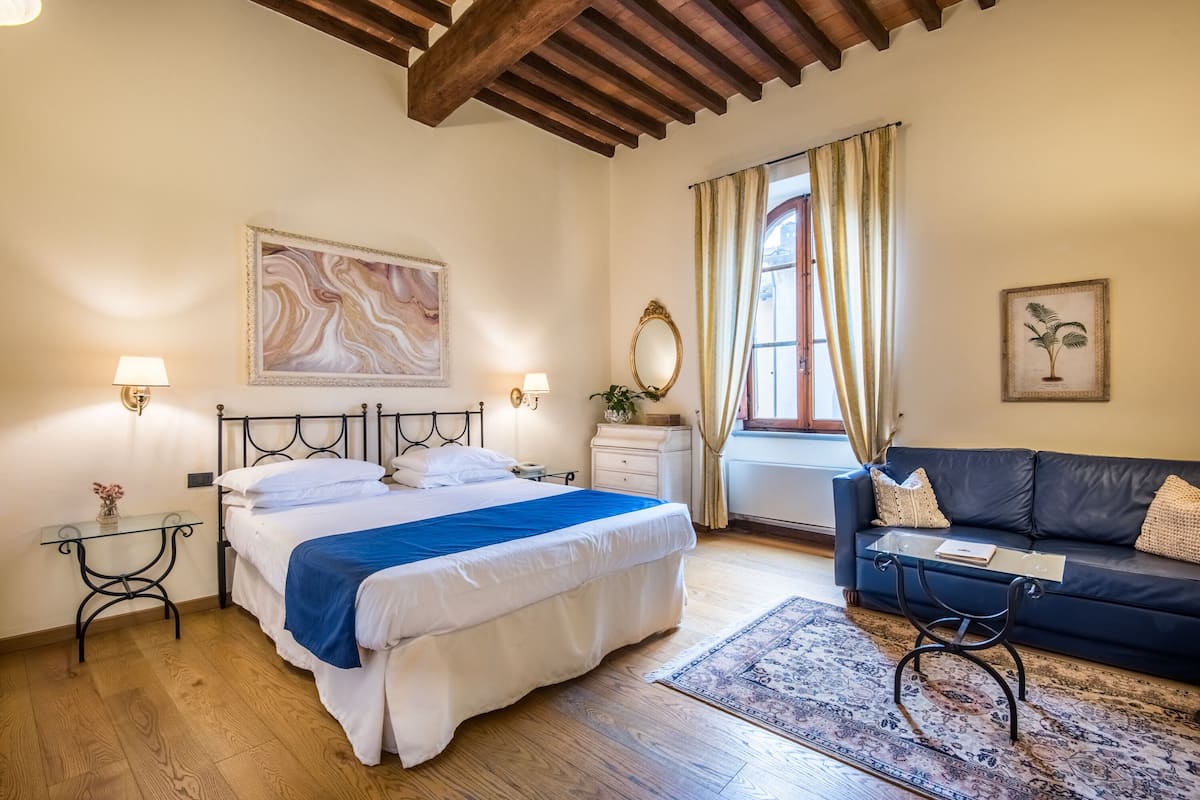 A bedroom with a bed, bedside tables, a sofa, a window with curtains, and a wall with a painting.
