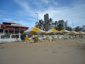 Uma praia com guarda-sóis amarelos e brancos, cadeiras de praia e um hotel ao fundo.