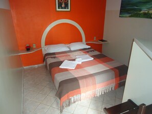 Uma cama com um cobertor xadrez e uma toalha dobrada, uma mesinha de cabeceira com um telefone, e um quadro na parede.
