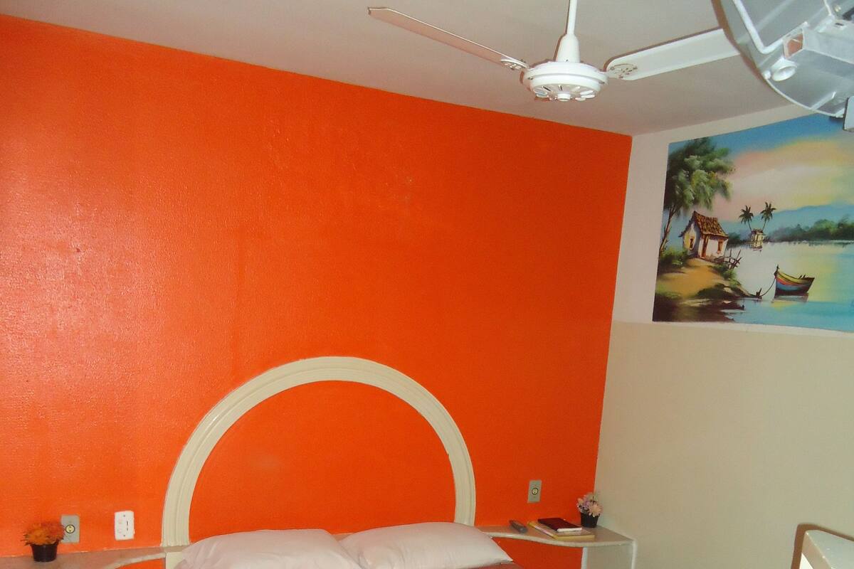 Ett sovrum med en orange vägg, en säng med ett rutigt täcke, en takfläkt och en tavla som föreställer en strandscen.
