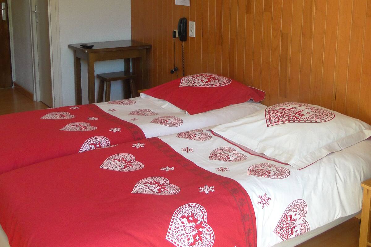 Une chambre d’hôtel avec un mur lambrissé en bois, un lit avec une literie rouge et blanche, deux lampes fixées au mur, un bureau en bois et une chaise.