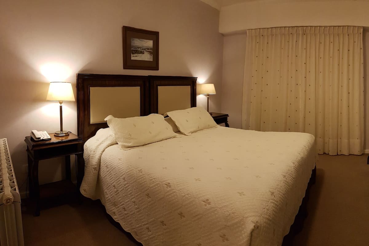 Habitación de hotel con una cama grande, dos mesitas de noche con lámparas, un escritorio con una silla, un televisor y una ventana con cortinas.