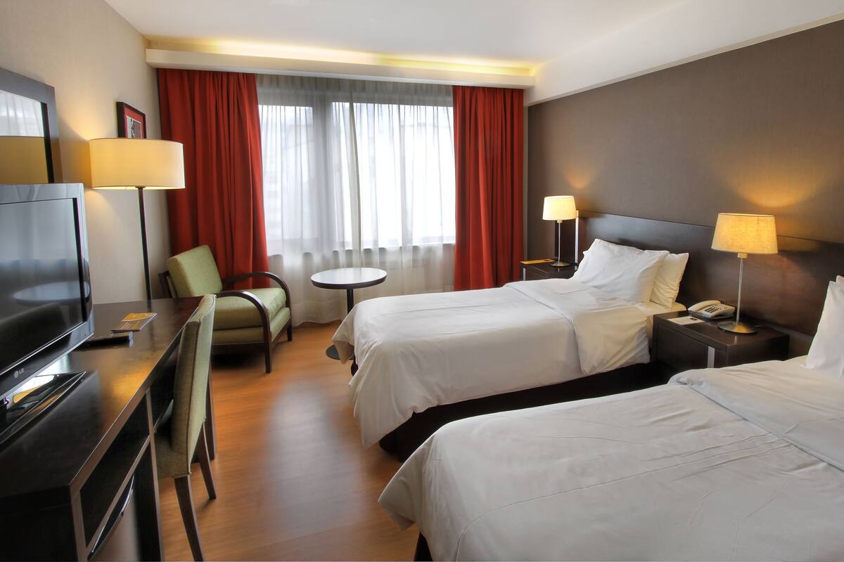 Une chambre d’hôtel avec deux lits, une télévision, un bureau, une chaise et une fenêtre avec des rideaux.