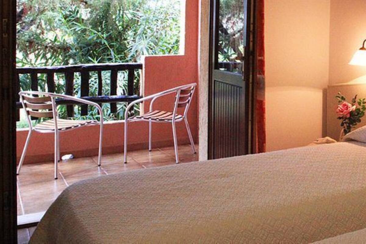 Habitación de hotel con dos camas, un balcón con una silla y una mesita de noche con lámpara.