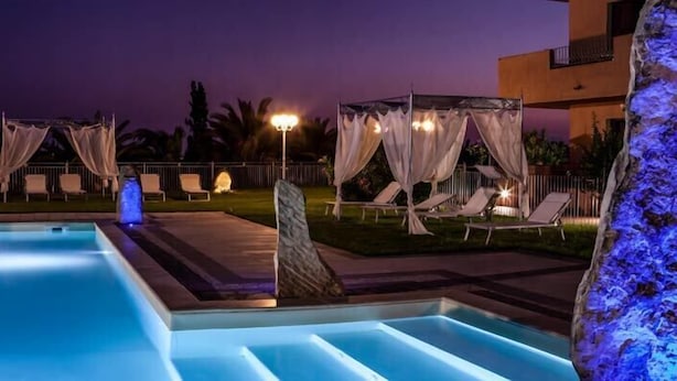A swimming pool with a night-time setting, including a well-lit pool area, lounge chairs, and a covered seating area with draped fabric. - I Ginepri (Quartucciu)