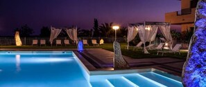 A swimming pool with a night-time setting, including a well-lit pool area, lounge chairs, and a covered seating area with draped fabric. - I Ginepri (Quartucciu)