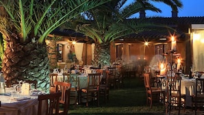 An outdoor dining area with tables and chairs, surrounded by palm trees and plants, under a covered patio. - I Ginepri (Quartucciu)