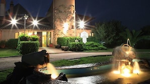 A well-lit hotel entrance with a fountain, a pathway, and a building with a tower. - I Ginepri (Quartucciu)