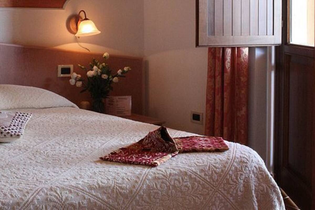 Una cama bien tendida con una almohada estampada y una colcha doblada, una lámpara de pared y un jarrón con flores.