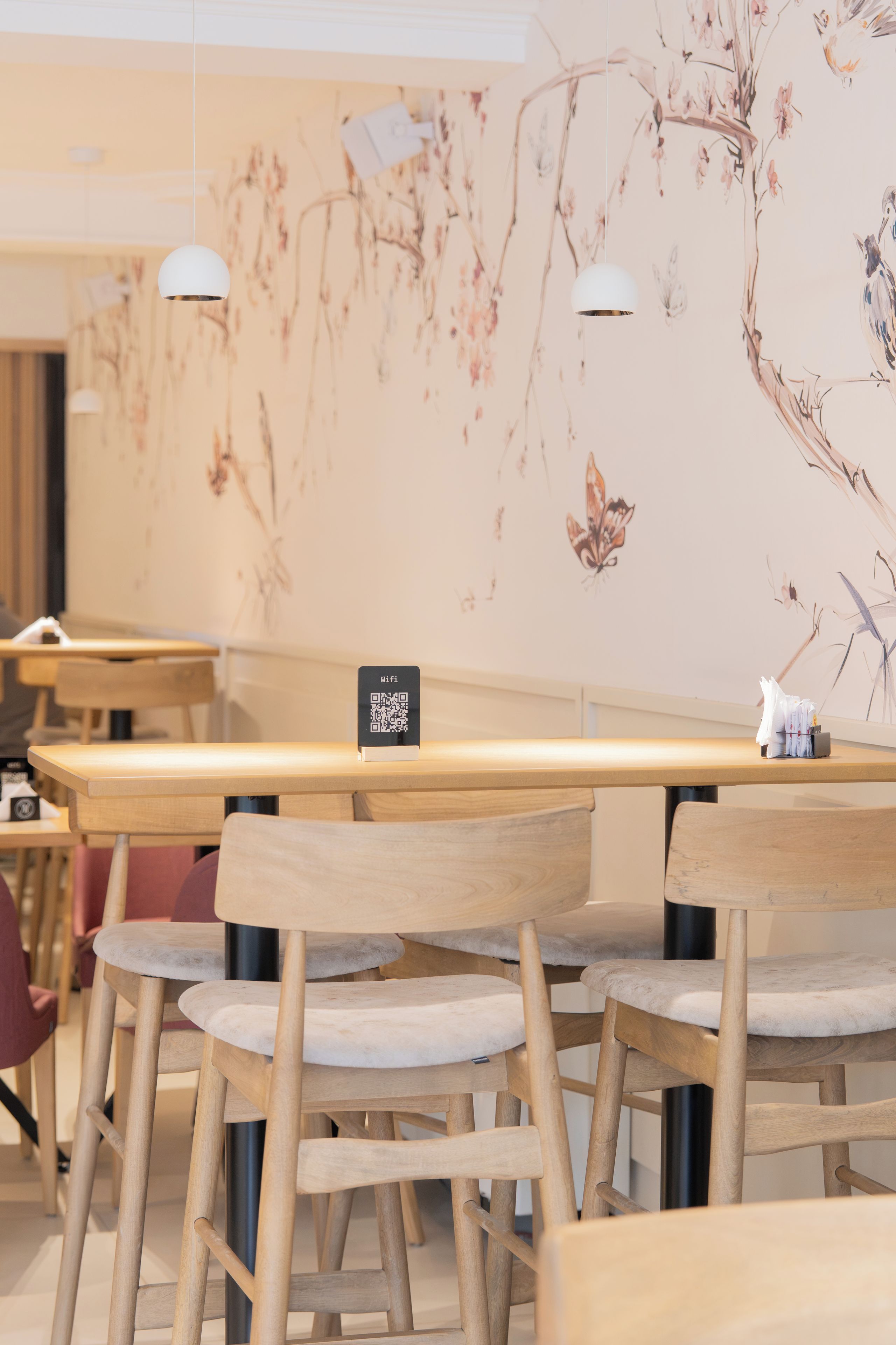 A cafe interior with wooden tables and chairs, a wall mural with floral and bird illustrations, and a sign indicating WiFi availability.