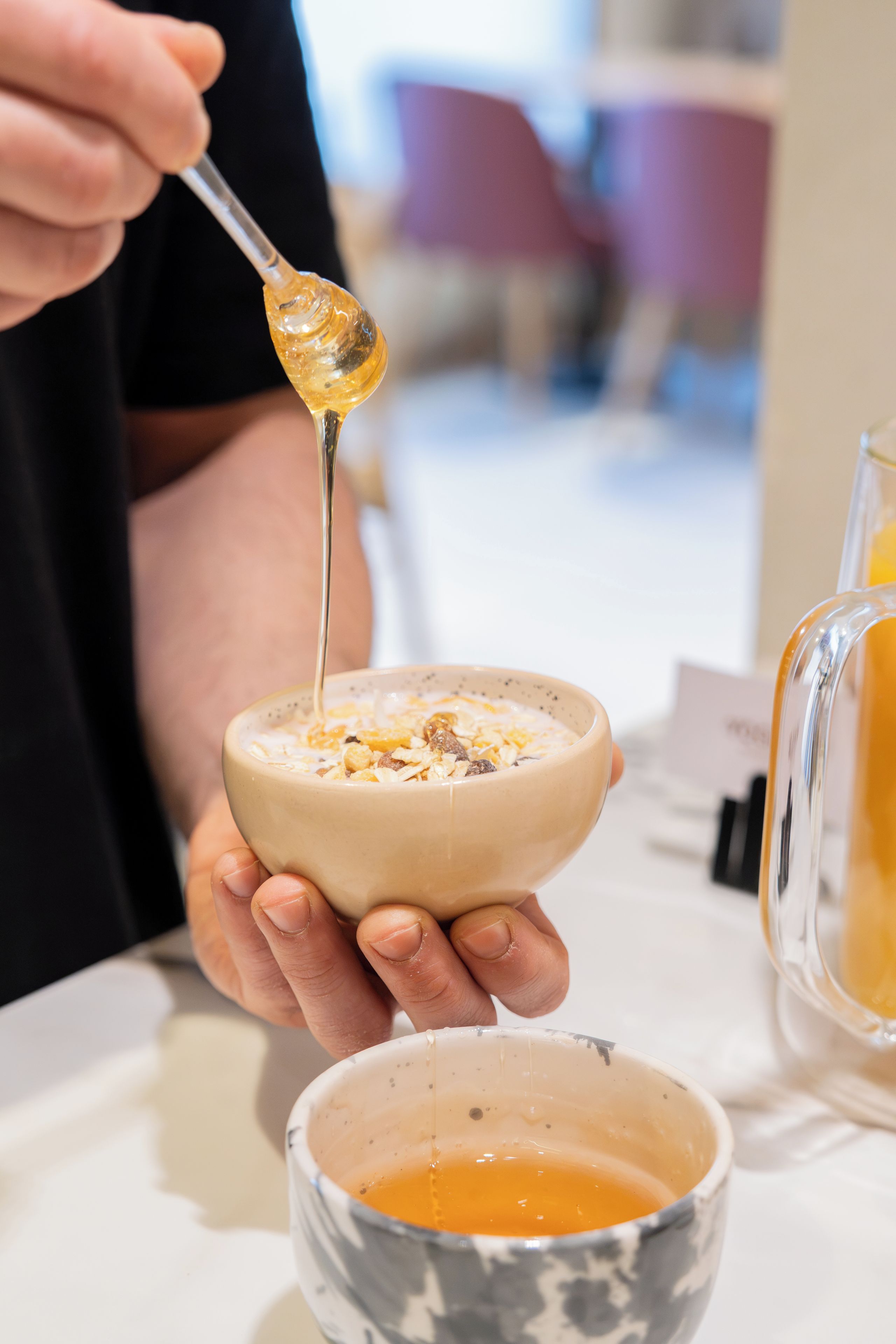 A person holding a bowl with a spoon, containing a liquid and granules, with a glass of orange juice on the table.