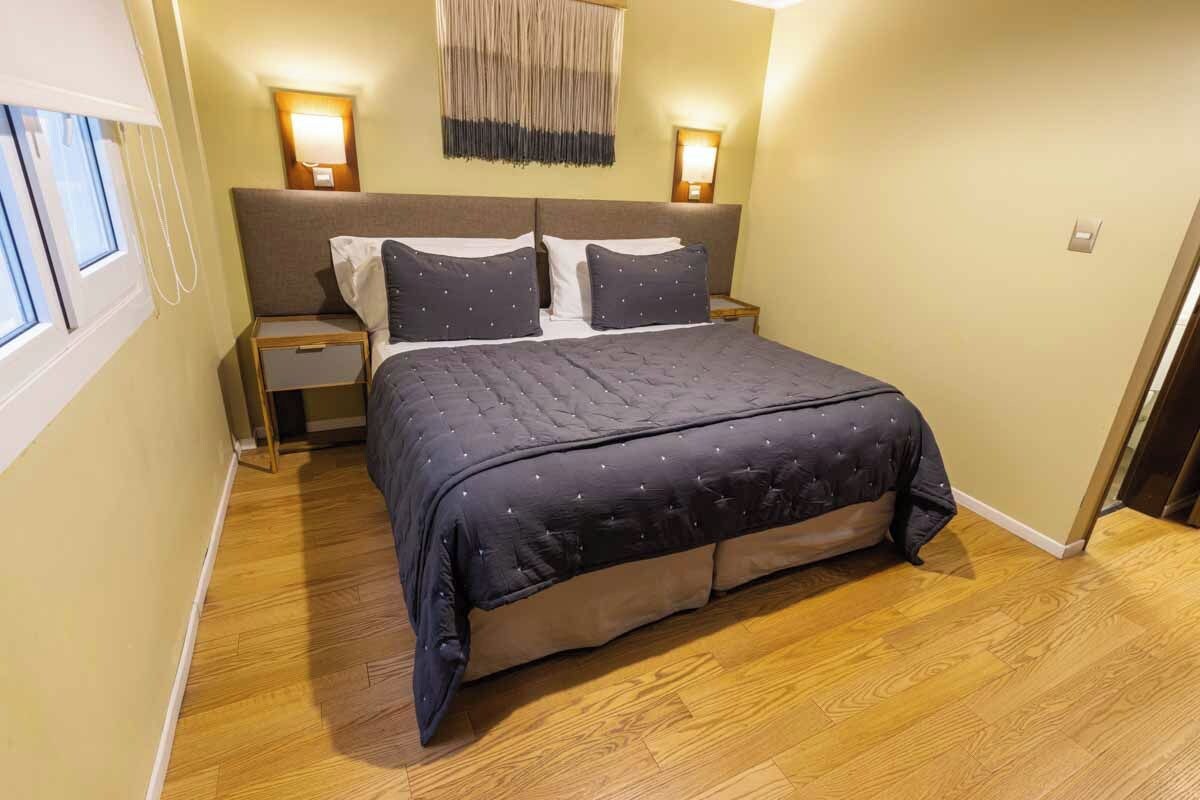 A bedroom with a bed, two bedside tables, a wall-mounted lamp, and a window.