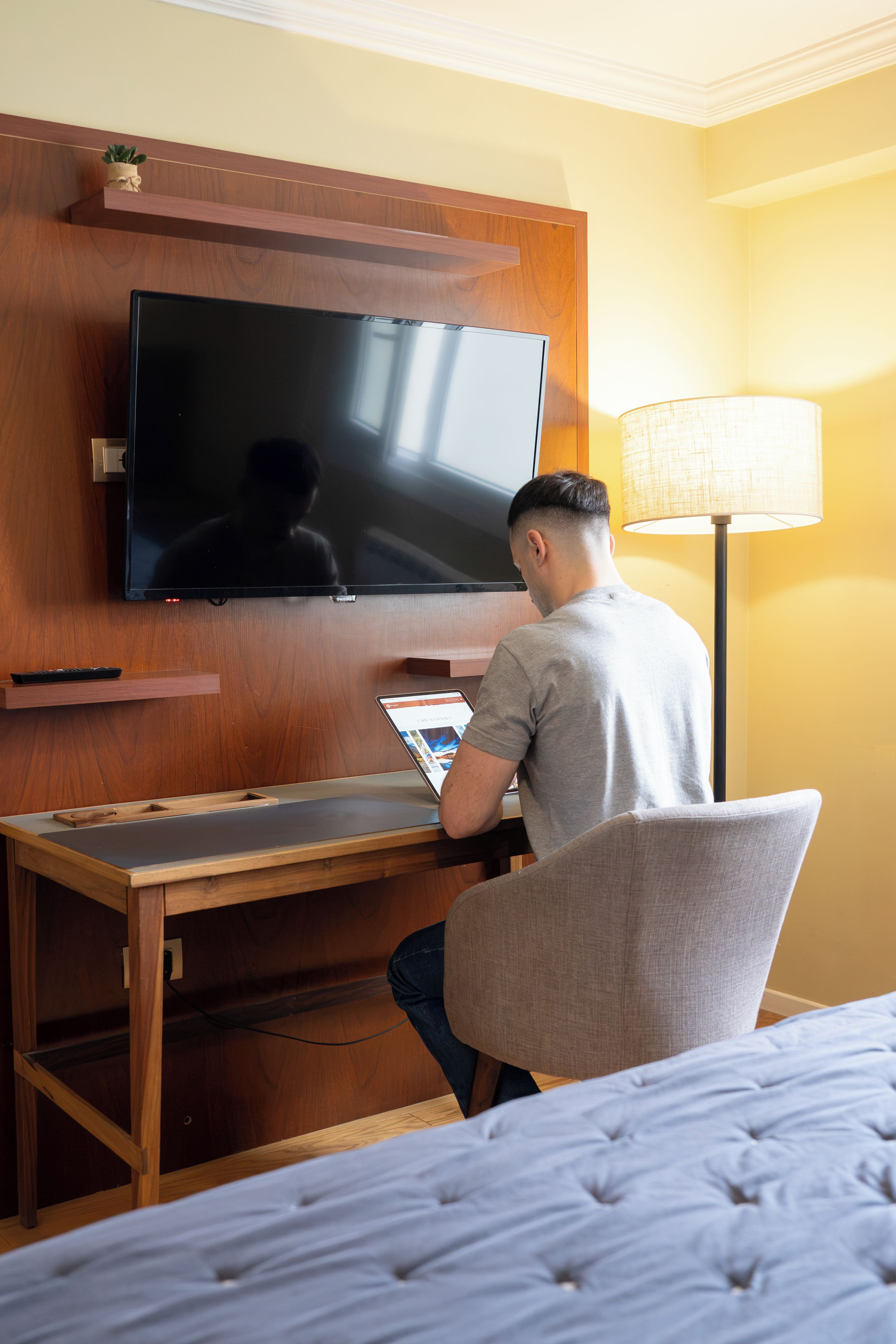 Et hotelværelse med et fladskærms-tv monteret på en trævæg, et skrivebord med en bærbar computer, en stol og en seng med en knaploftsgavl.