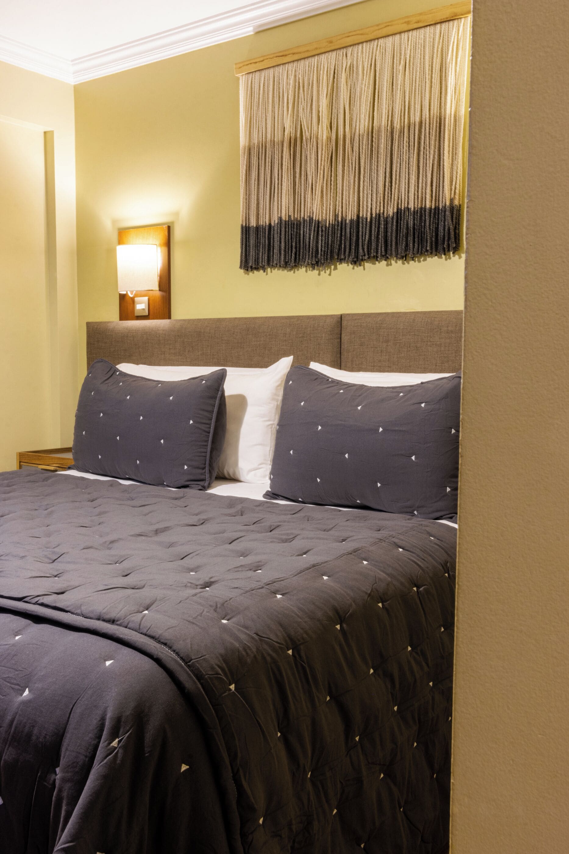 Een net opgemaakt bed met een donkere, gewafelde sprei en twee kussens met een subtiel patroon. Boven het bed is een wandlamp te zien.