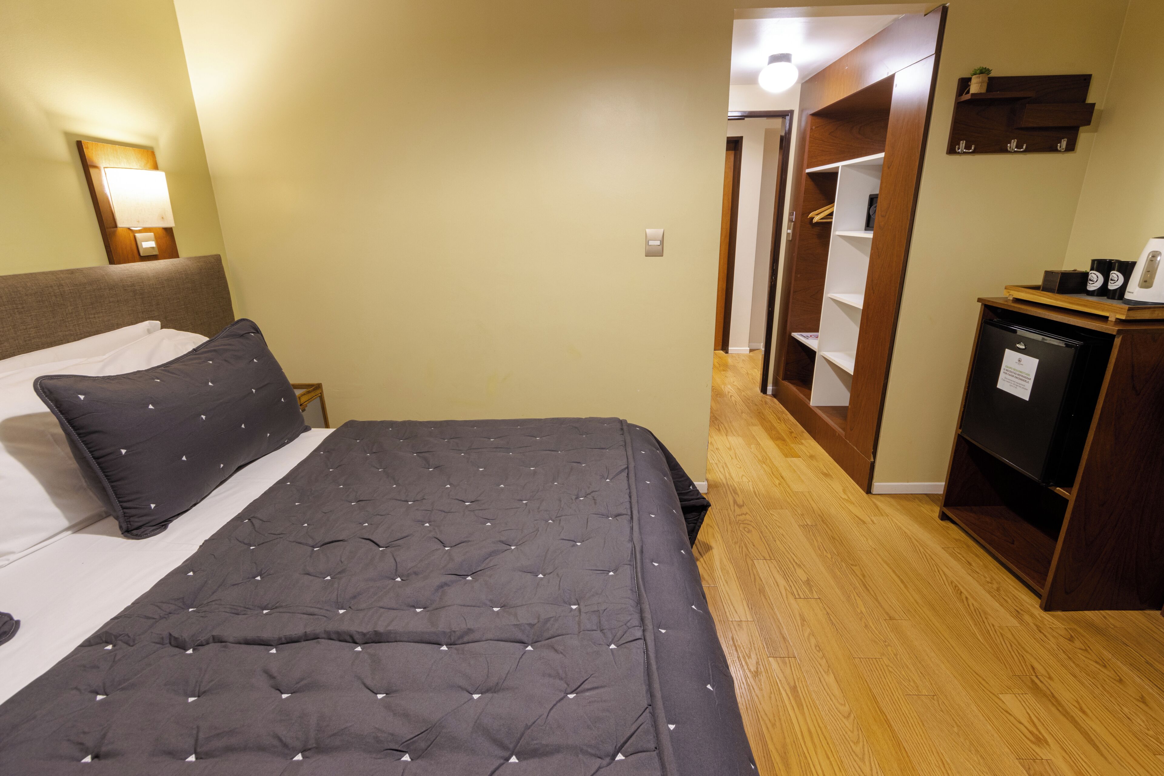 A hotel room with a bed, a nightstand, a desk with a computer, and a wardrobe.