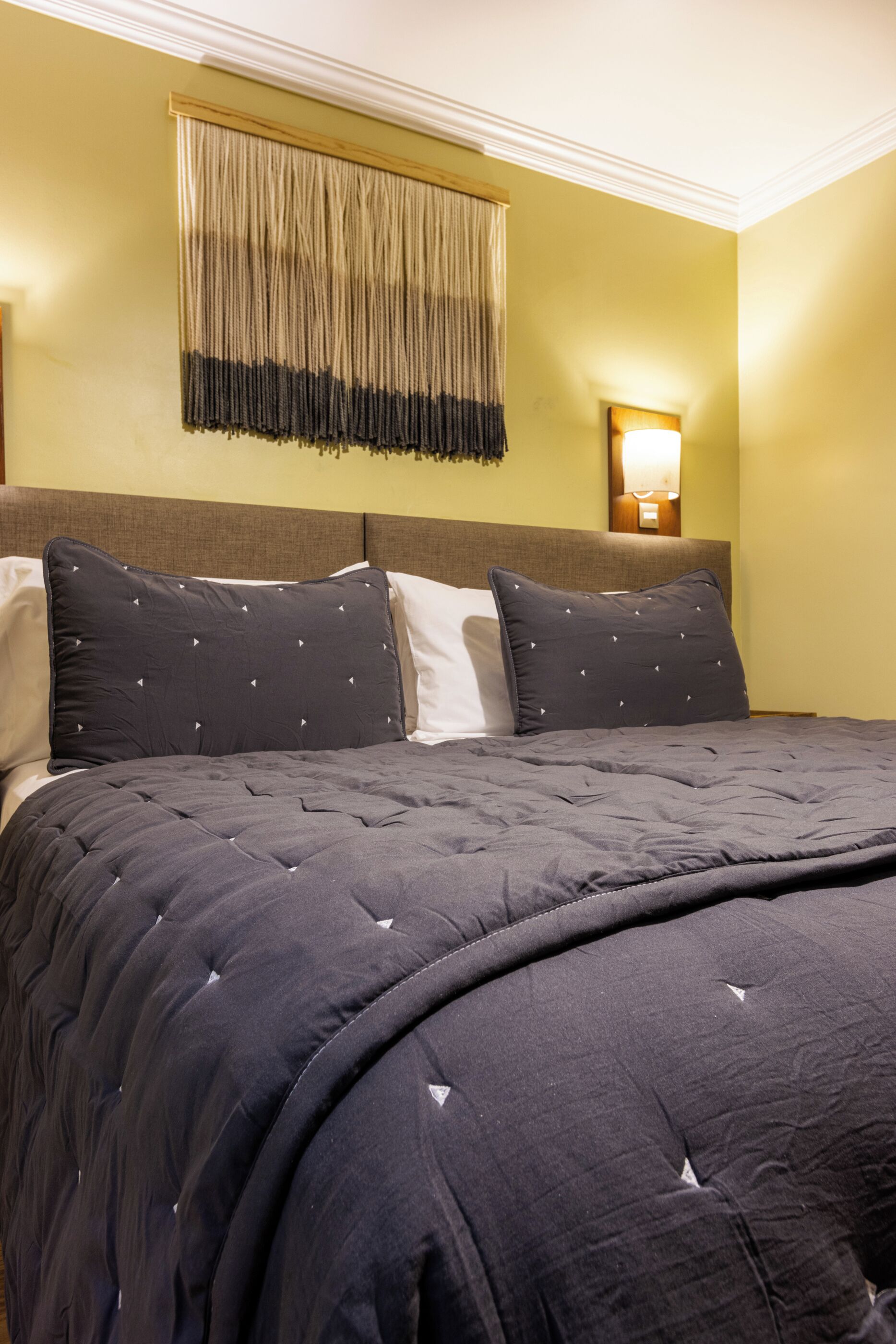 A neatly made bed with a dark, tufted bedspread and two pillows, a wall-mounted light fixture, and a textured curtain.