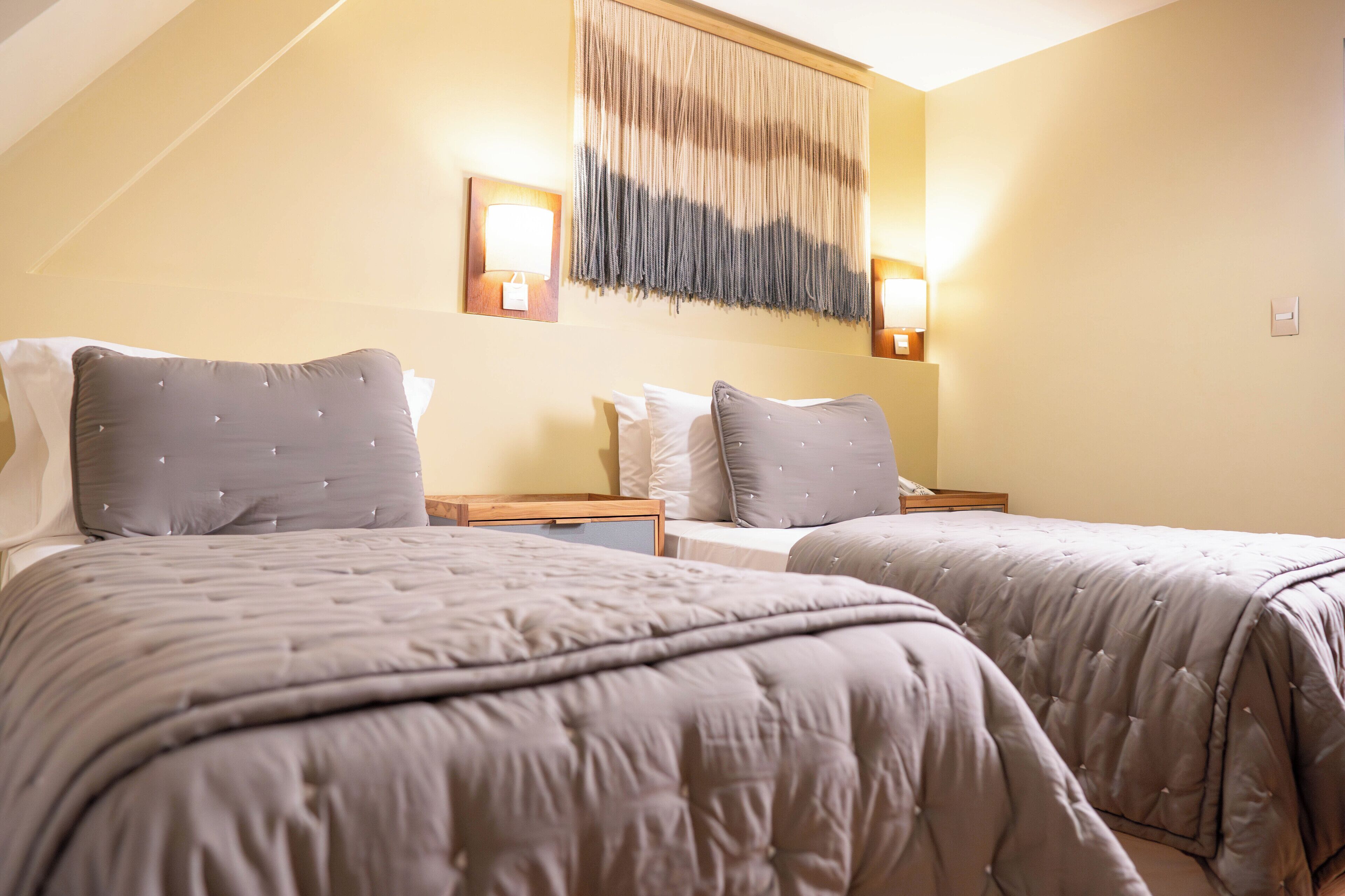Habitación de hotel con dos camas, cada una con edredón gris acolchado y almohadas, una mesita de noche de madera y lámparas fijadas a la pared.