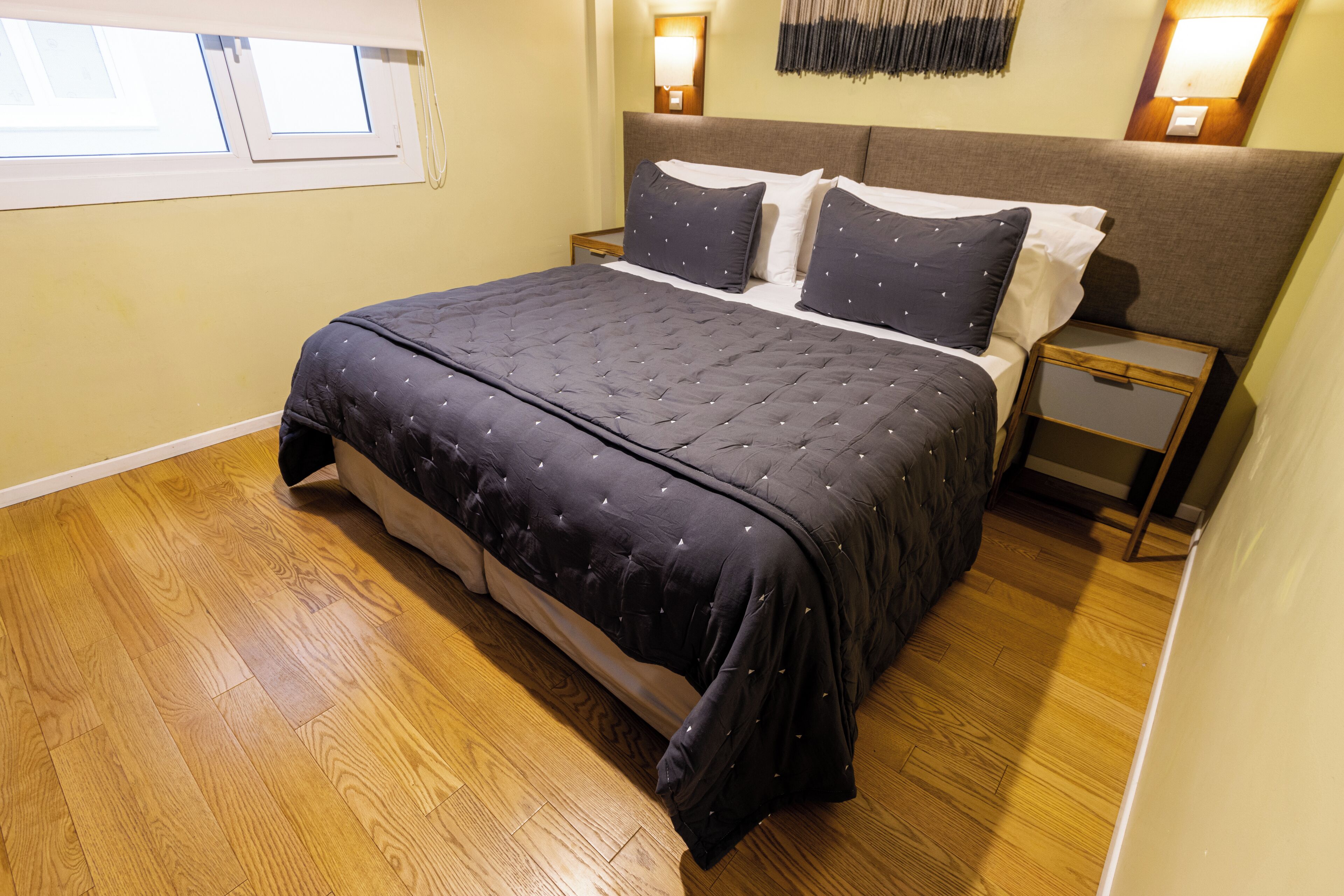 A neatly made bed with a dark quilt, wooden flooring, and bedside lamps.