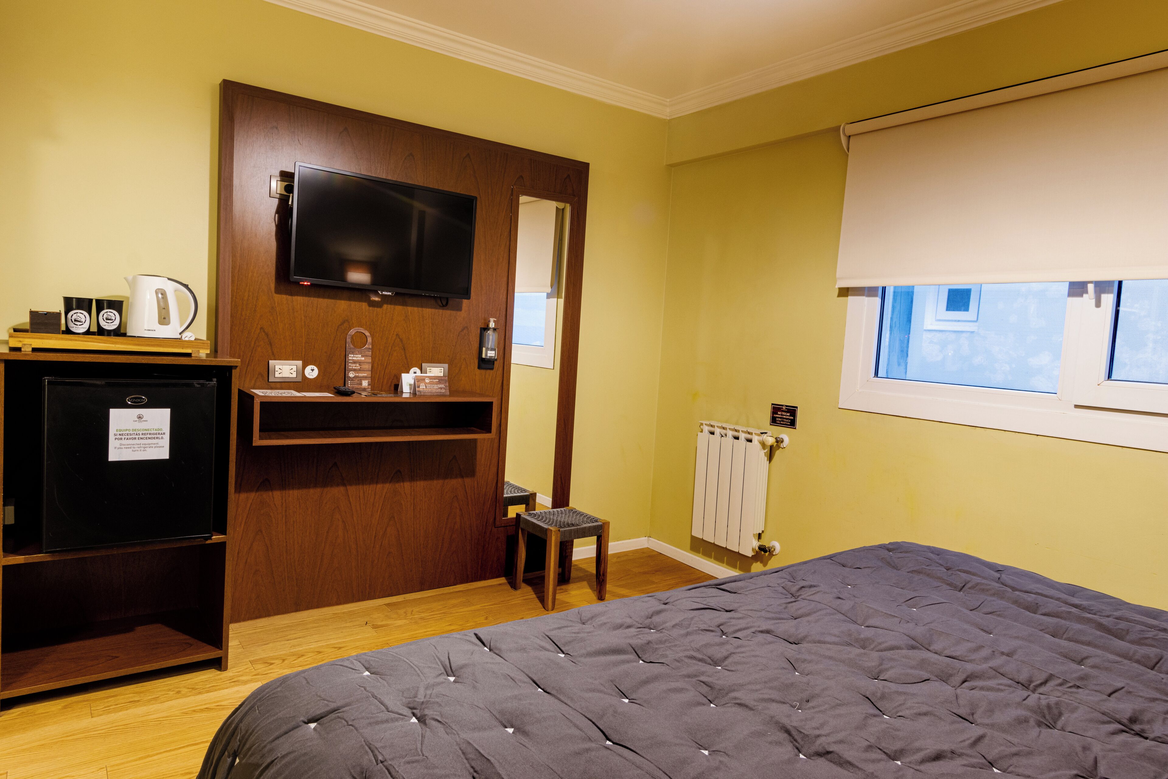 Habitación de hotel con una cama, un televisor montado en un mueble de madera, una mesita con una tetera y una ventana con vistas al exterior.