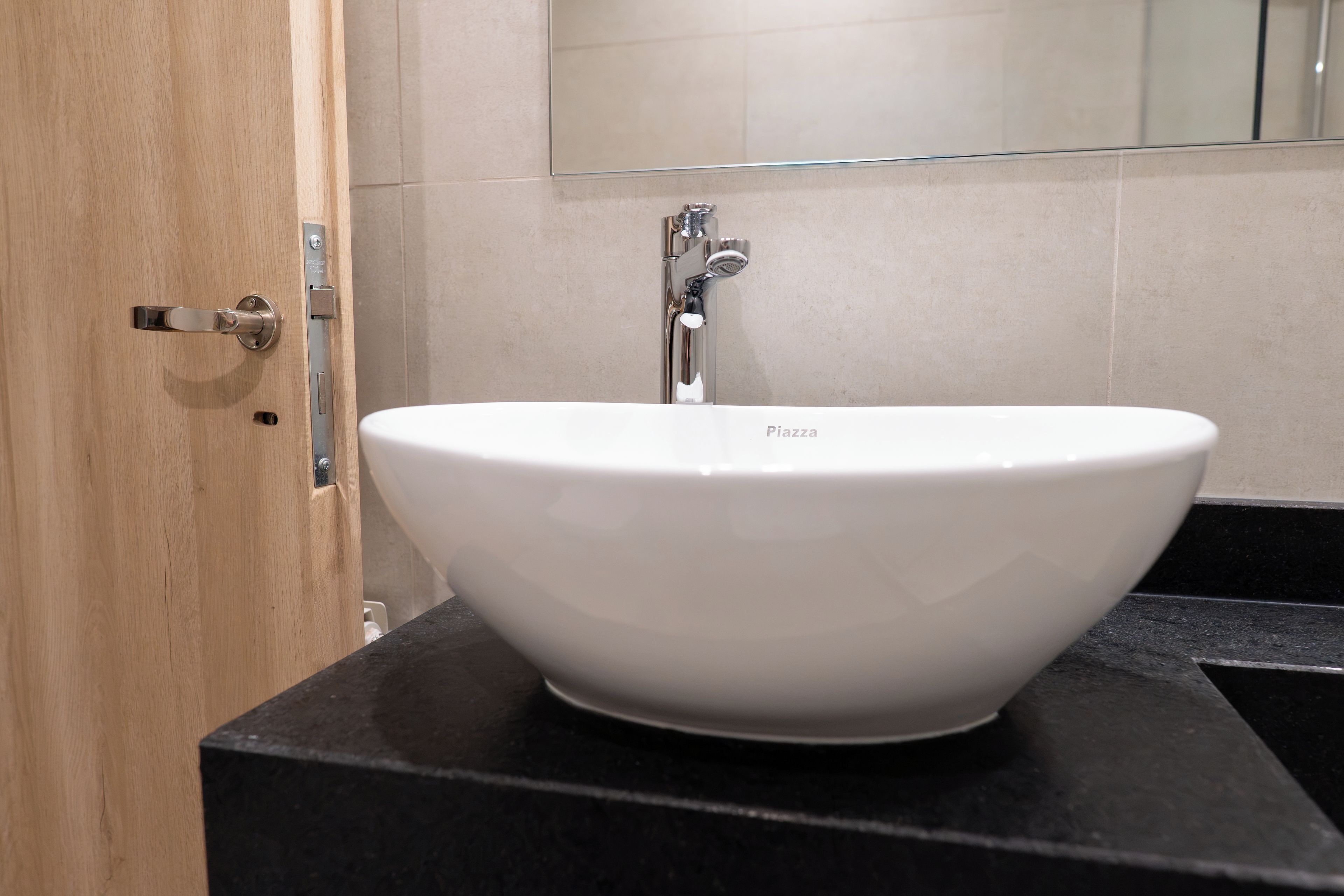 A white vessel sink with a faucet on a black countertop, a wooden door with a silver handle, and a mirror above the sink.