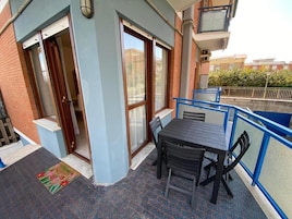Un área de estar al aire libre con una mesa y sillas, una barandilla azul y una puerta que da a un espacio interior.