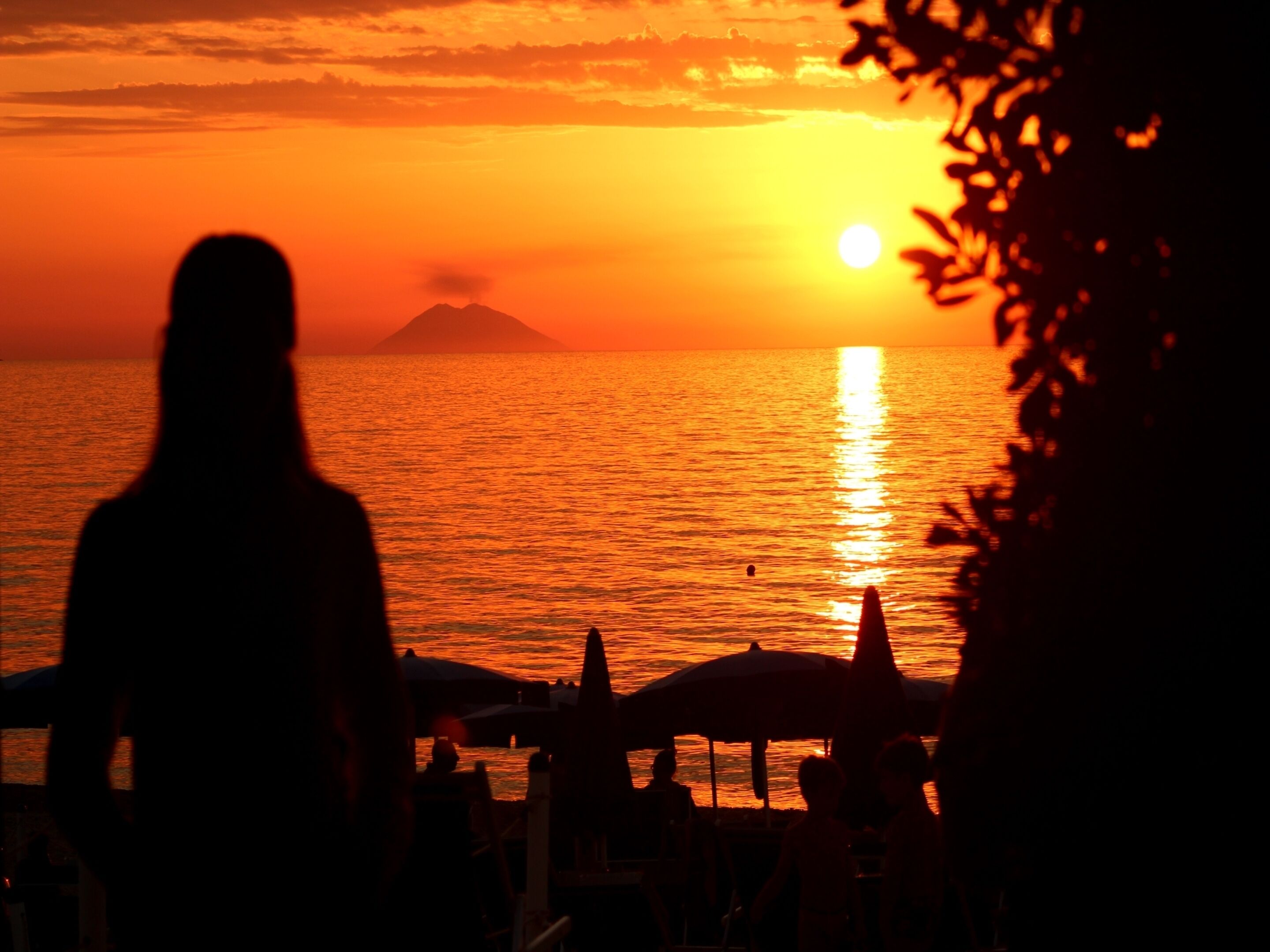 En silhuet af en person, der ser ud over en solnedgang over havet, med en fjerntliggende bjergkæde og strandparasoller i det fjerne.