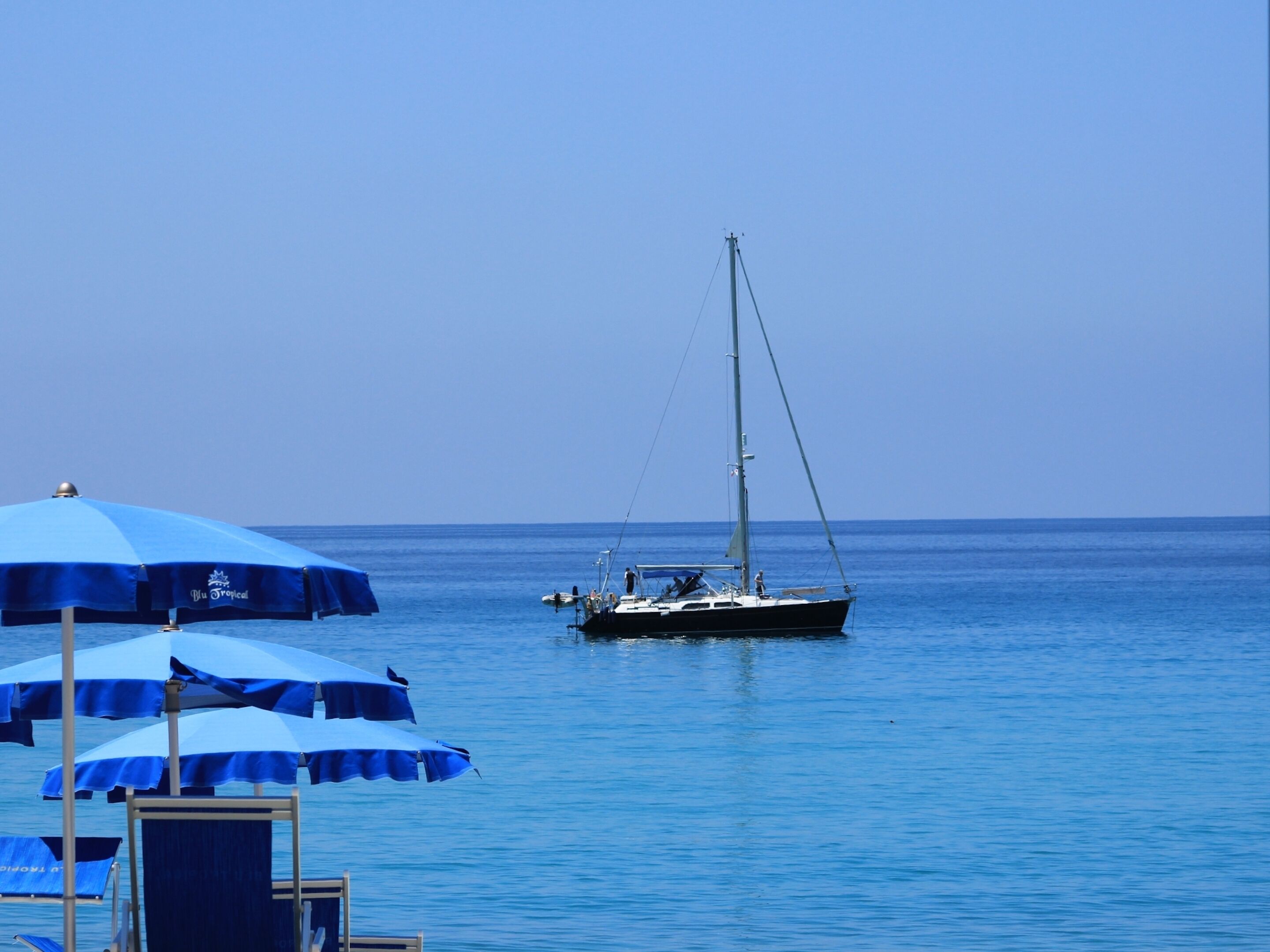 Ein Segelboot auf ruhigem Wasser mit blauen Sonnenschirmen und Liegestühlen am Ufer.