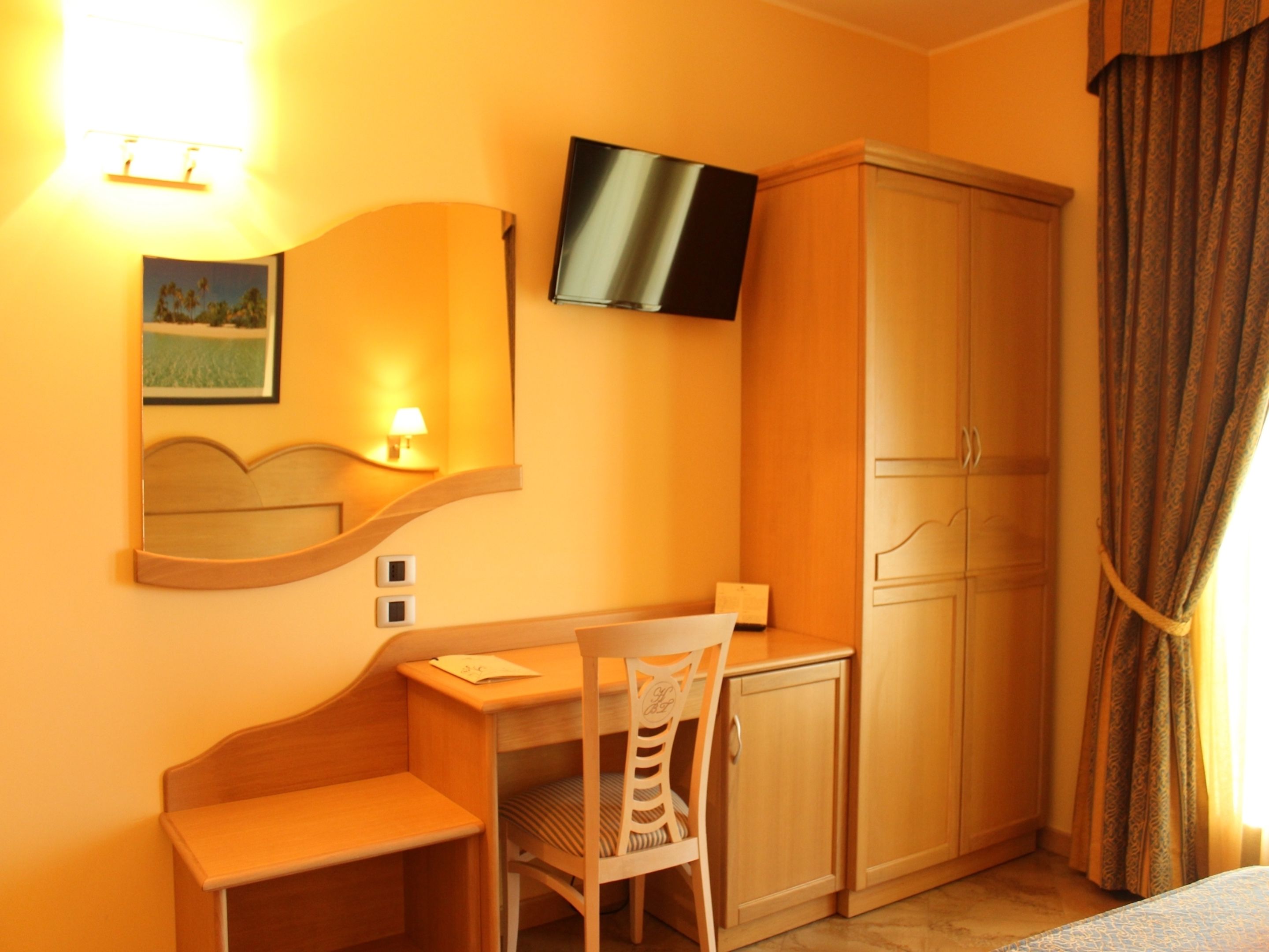 Une chambre d’hôtel avec un bureau, une chaise et une armoire en bois. Un téléviseur est fixé au mur et une lampe se trouve sur le bureau.